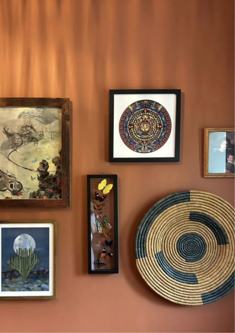 Gallery wall with framed artwork and decorations on a brown wall, including a colorful circular design, a frame with a butterfly display, a painting of a cactus and moon, and other framed pictures.