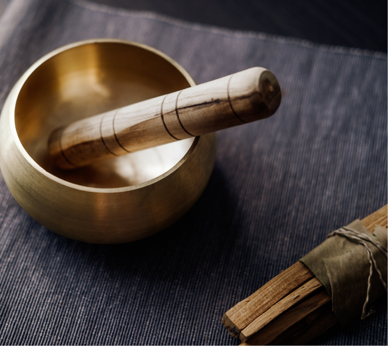Close-up of a singing bowl with a wooden striker inside, and a bundle of sticks wrapped with string nearby, placed on a dark fabric surface.