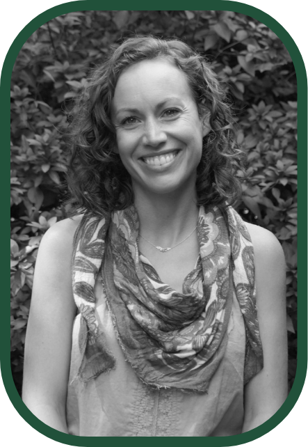 A smiling woman with curly hair wearing a scarf and sleeveless top, standing outdoors in front of leafy bushes.