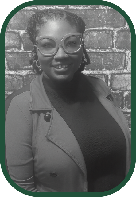 A smiling woman with glasses and braids, wearing a jacket, standing in front of a brick wall.
