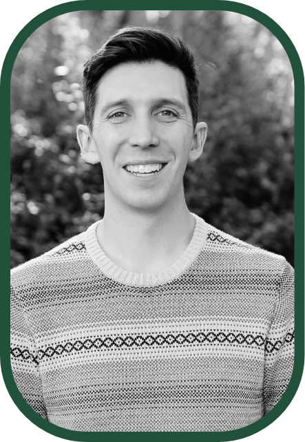 Black and white portrait of a smiling young man with short dark hair, wearing a patterned sweater, standing outdoors with blurred trees in the background.