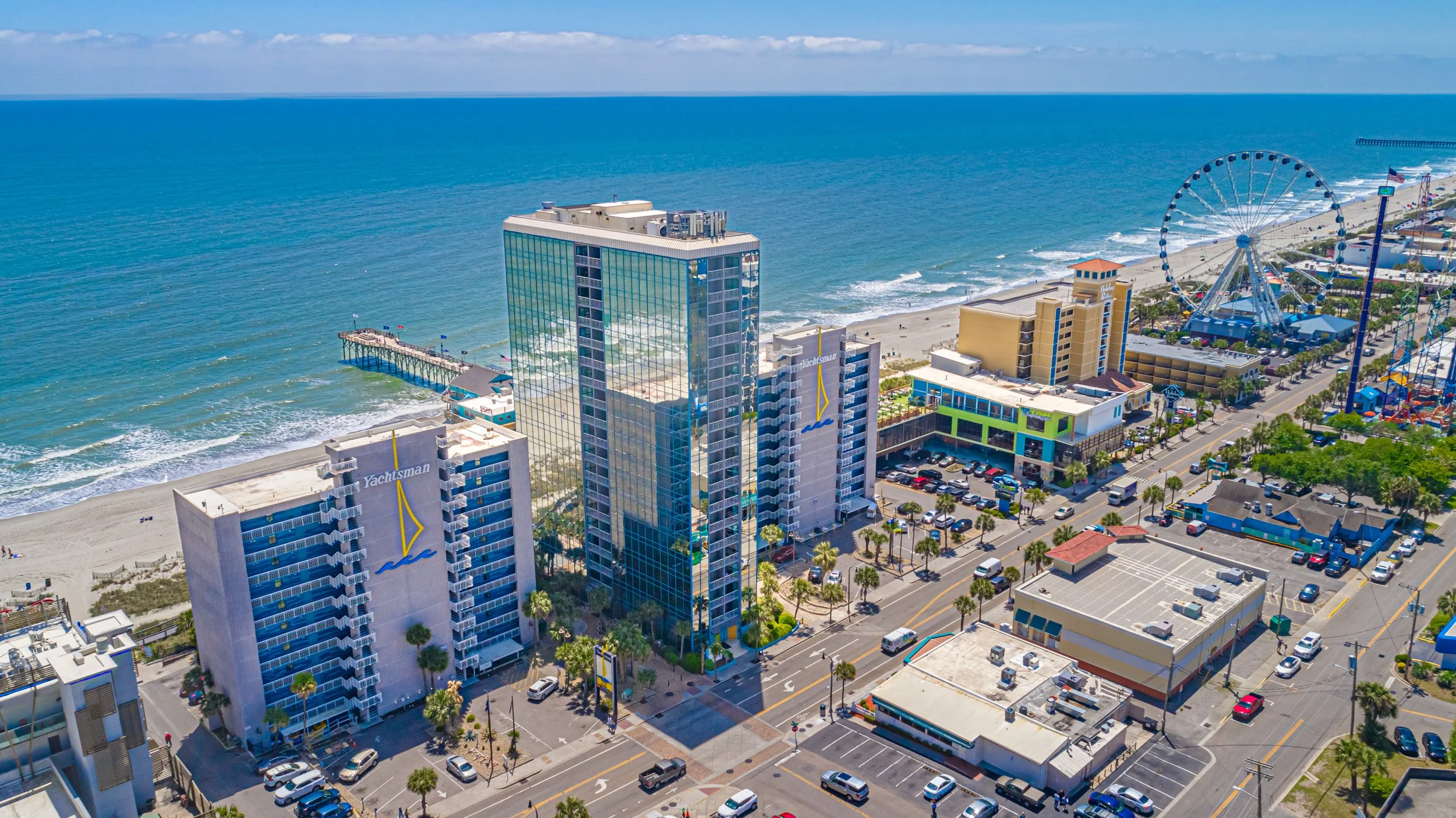 Aerial view of a beachfront amusement park with a Ferris wheel, high-rise hotels, parking lot, and the ocean in the background on a sunny day.