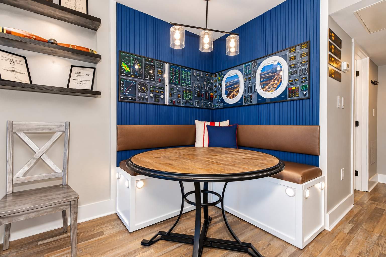 Corner seating with a round wooden table in a room with aviation-themed decor, including a wall display of airplane instrument panel images and airplane windows, wood flooring, and a mix of modern and vintage furniture.