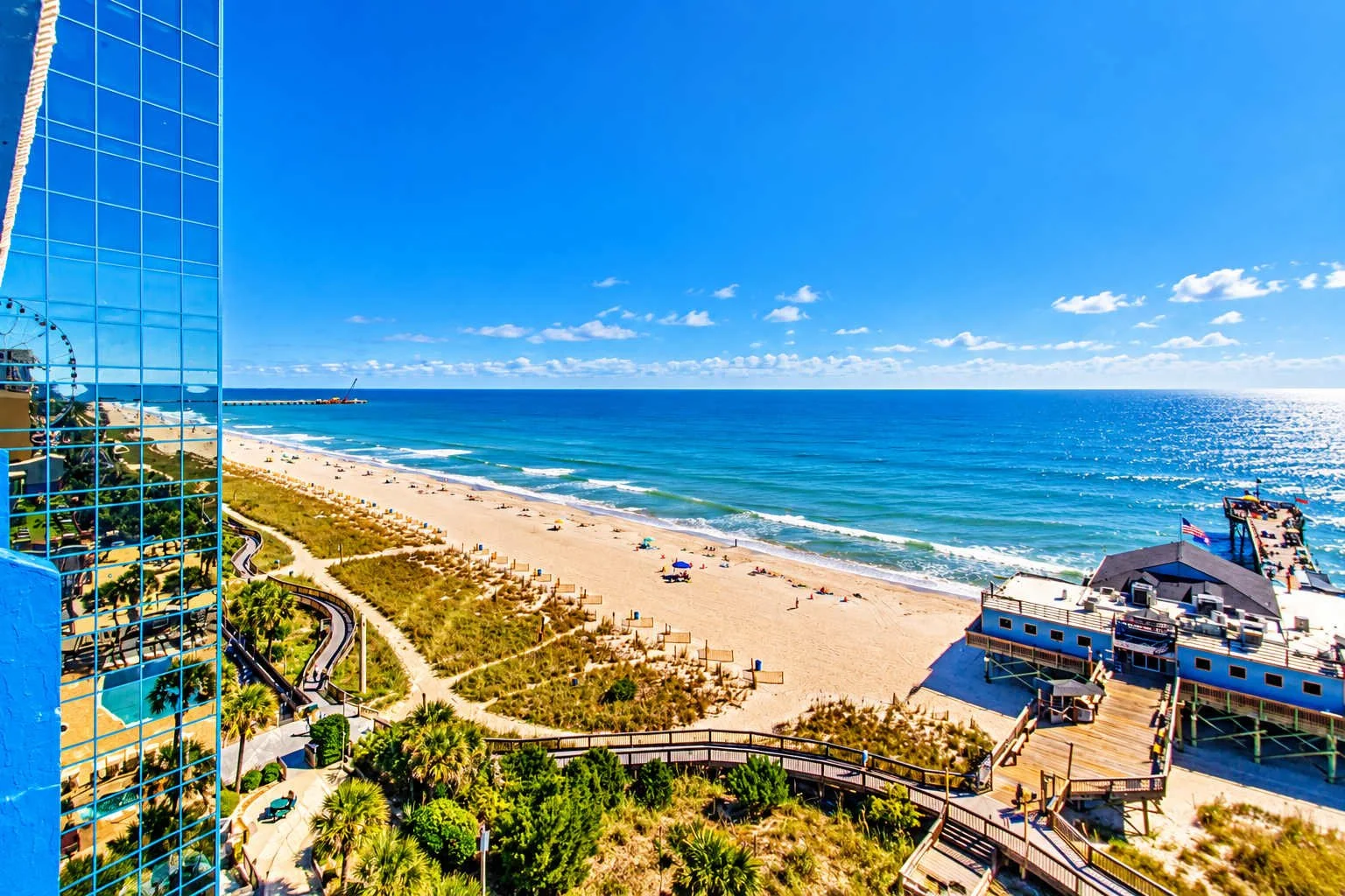A view of a beach with blue water, sandy shore, and a clear sky, with buildings and a boardwalk along the coast.