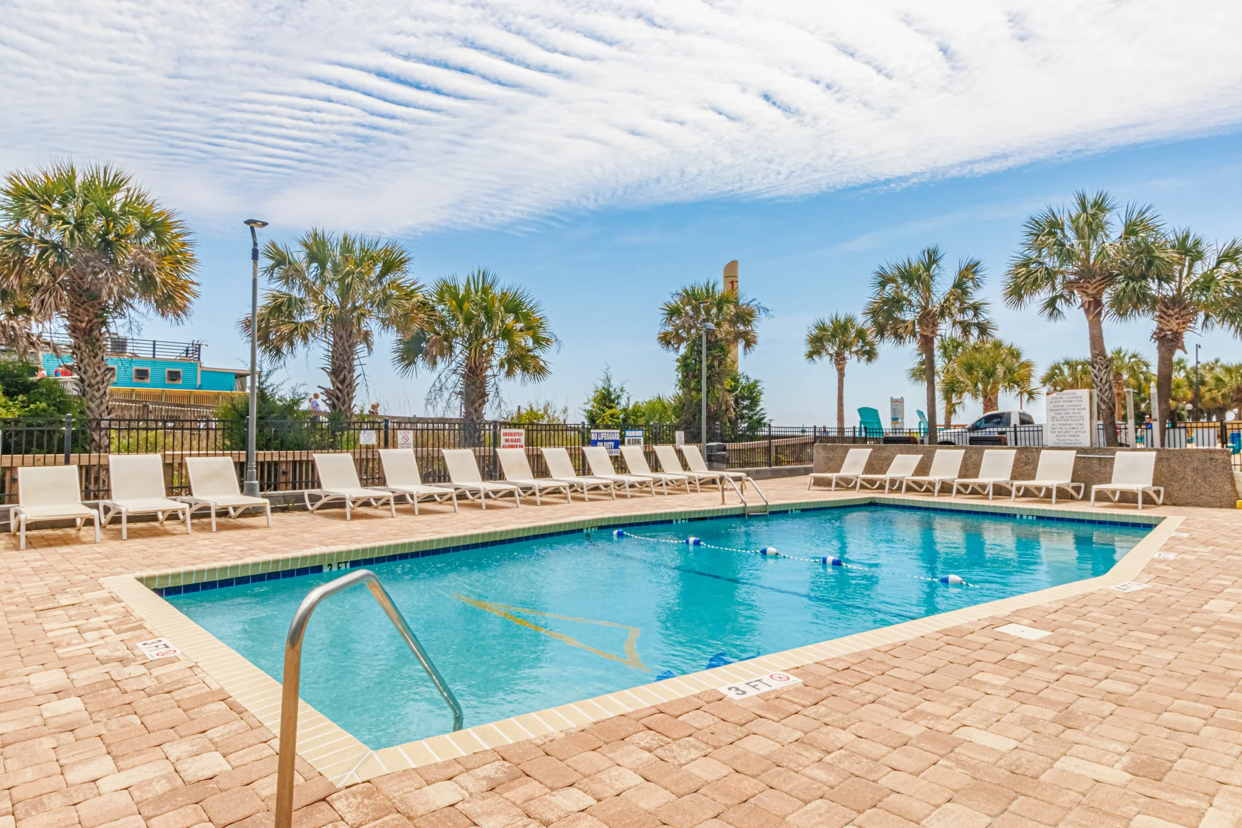Outdoor swimming pool with deck chairs, surrounded by palm trees and a black metal fence. There are clear blue skies with some clouds.