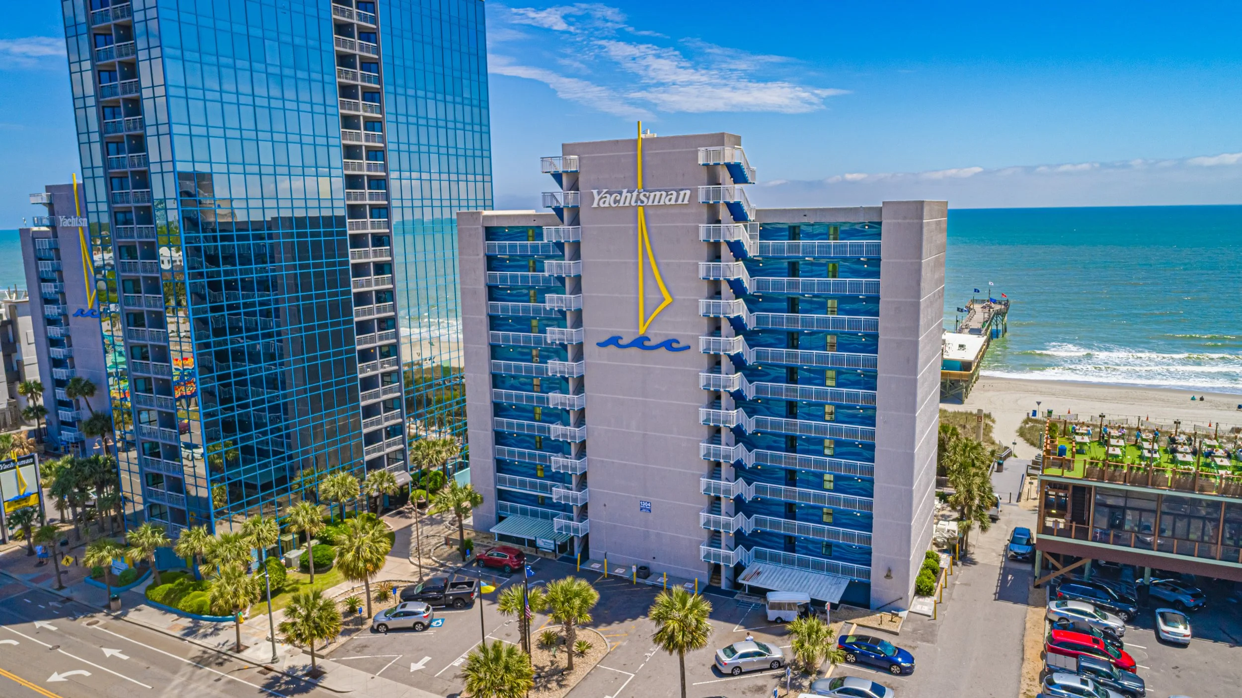Aerial view of a beachfront hotel called Yachtsman with a large glass-tiled building reflecting the sky and ocean, balconies on the side, palm trees, and a parking lot with cars. The beach and ocean are visible in the background.