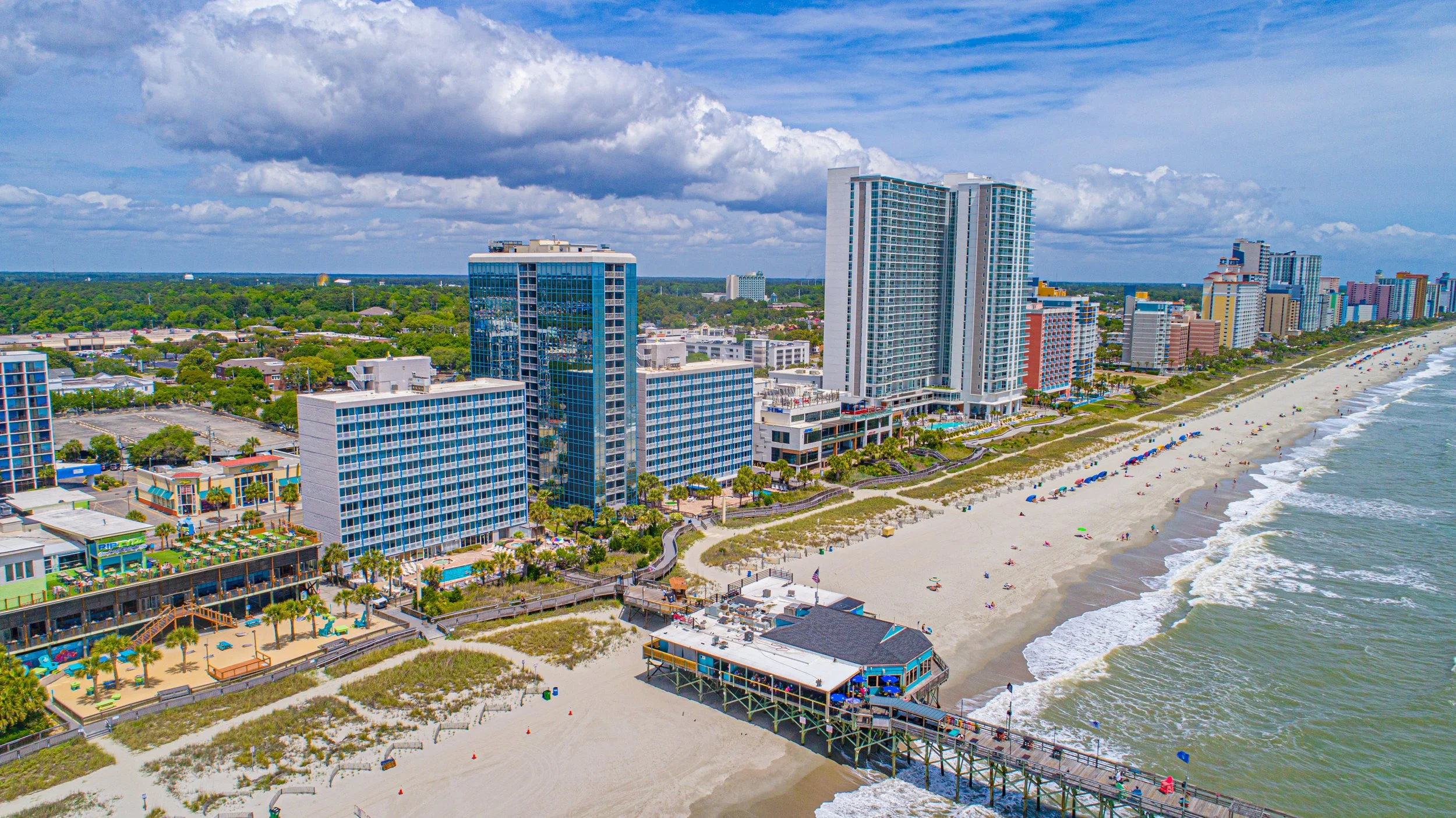 Aerial view of a beach city with high-rise buildings along the shoreline, sandy beach with umbrellas, and ocean waves, under partly cloudy skies.