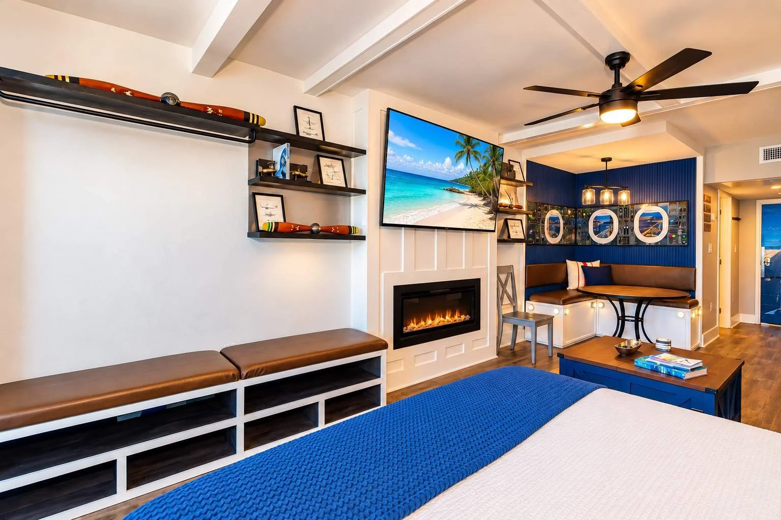 Living room with flat-screen TV displaying a beach scene, electric fireplace, blue accent wall with round mirrors, seating area with table, and tropical decor.