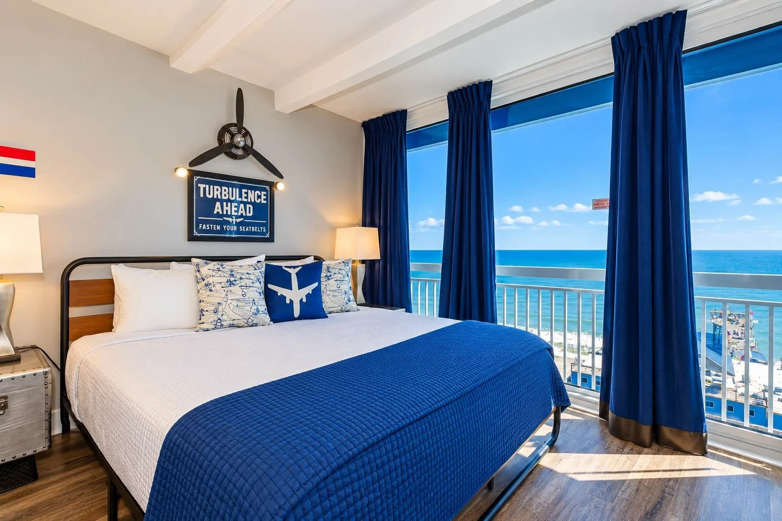 Beachside hotel room with a large bed, nautical-themed pillows, blue curtains, and ocean view from a balcony.