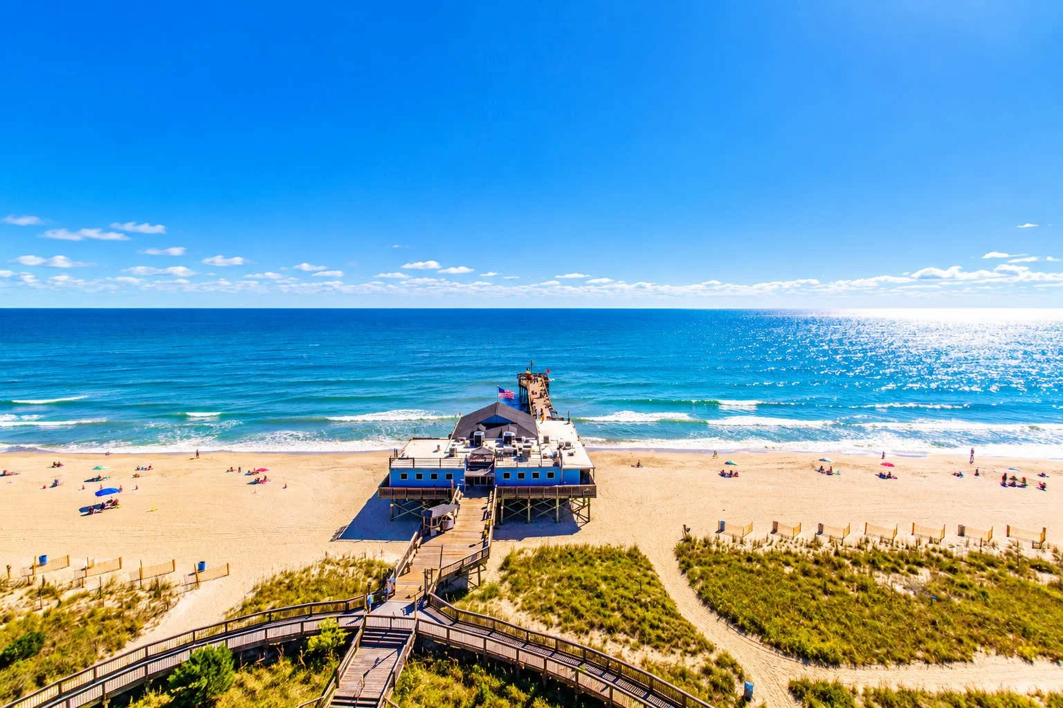 Aerial view of a pier extending into the ocean from a sandy beach with colorful umbrellas, green dunes, and a clear blue sky.