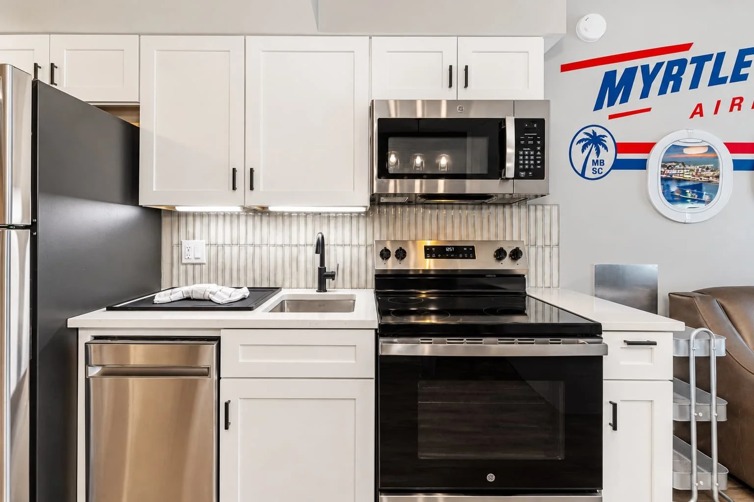 Modern kitchen with white cabinets, stainless steel appliances including microwave and oven, and a black refrigerator. Behind the sink area, there is a backsplash with vertical tiles. To the right, a wall features a large mural of Myrtle Beach, South