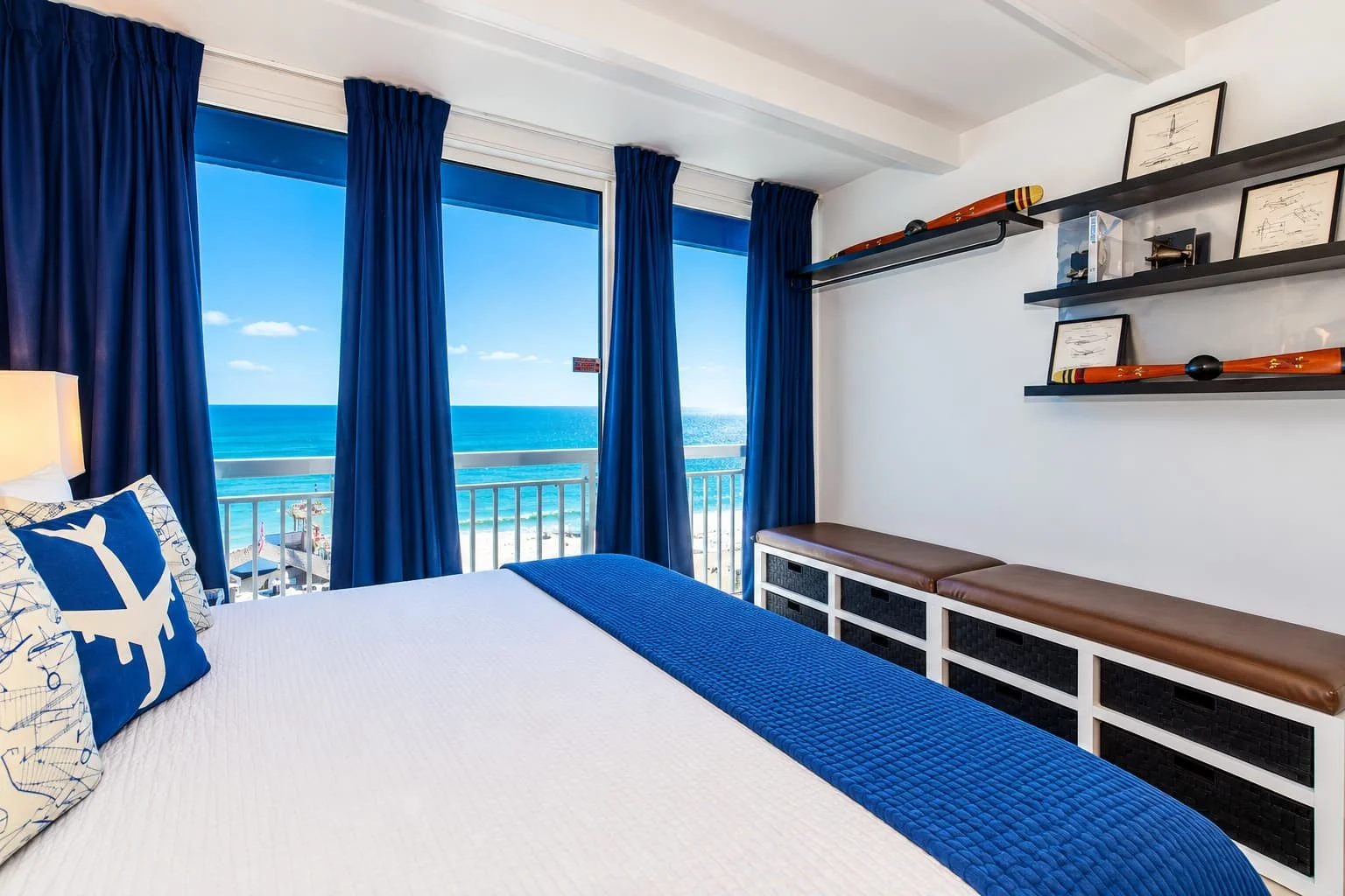 Hotel room with a view of the ocean, featuring a bed with blue and white decor, dark blue curtains, and seaside-themed decorations on the wall shelves.