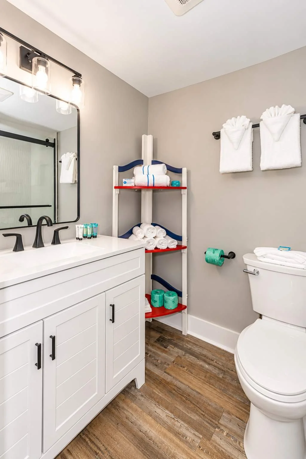 Bathroom with white vanity, black fixtures, mirror, shelves with towels and toiletries, two towel towels on a black rod, toilet with a towel on top, wood flooring, and a towel roll on the wall.