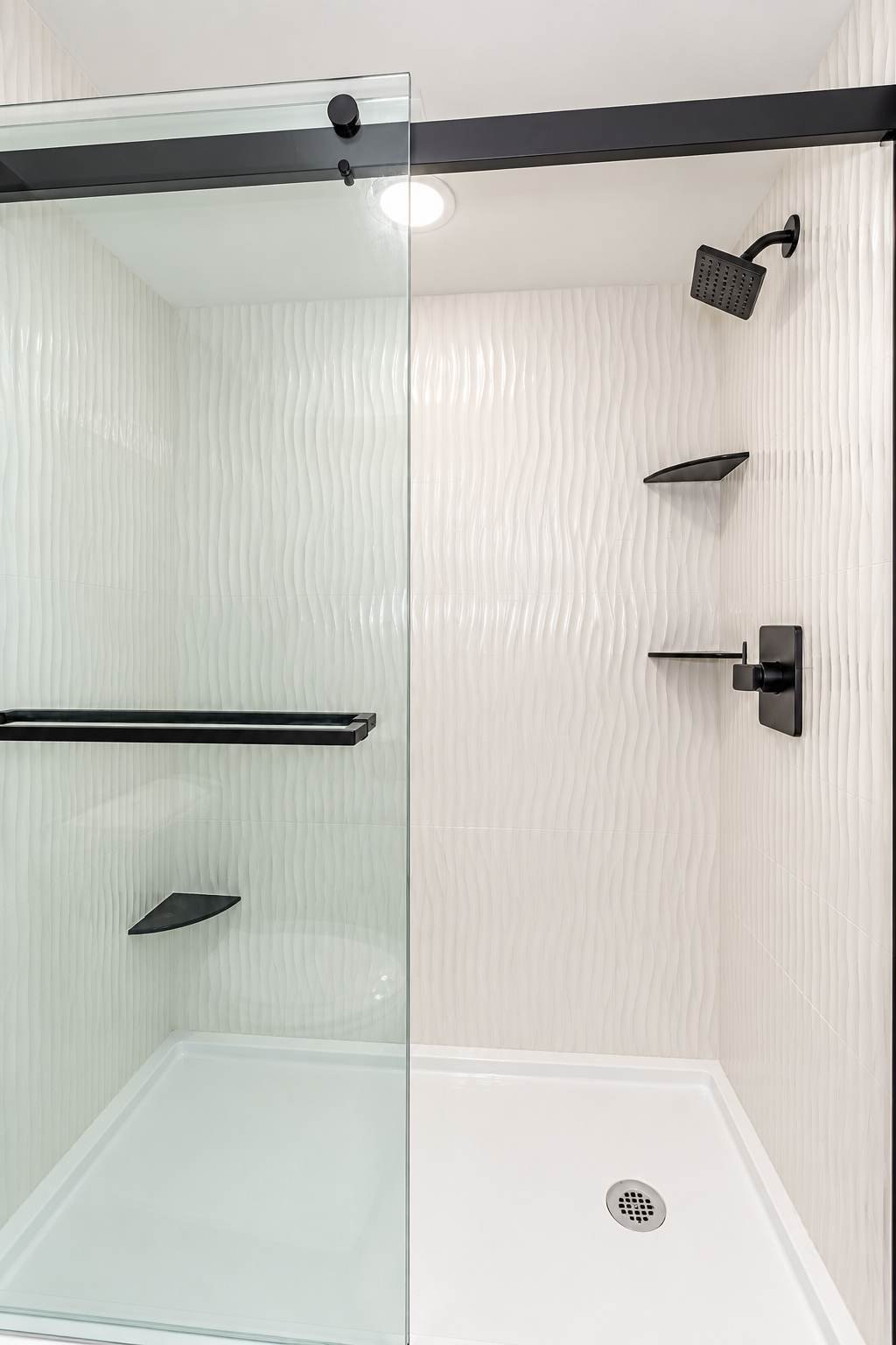 Modern shower with white textured walls, black fixtures, a glass door, and a rainfall showerhead.