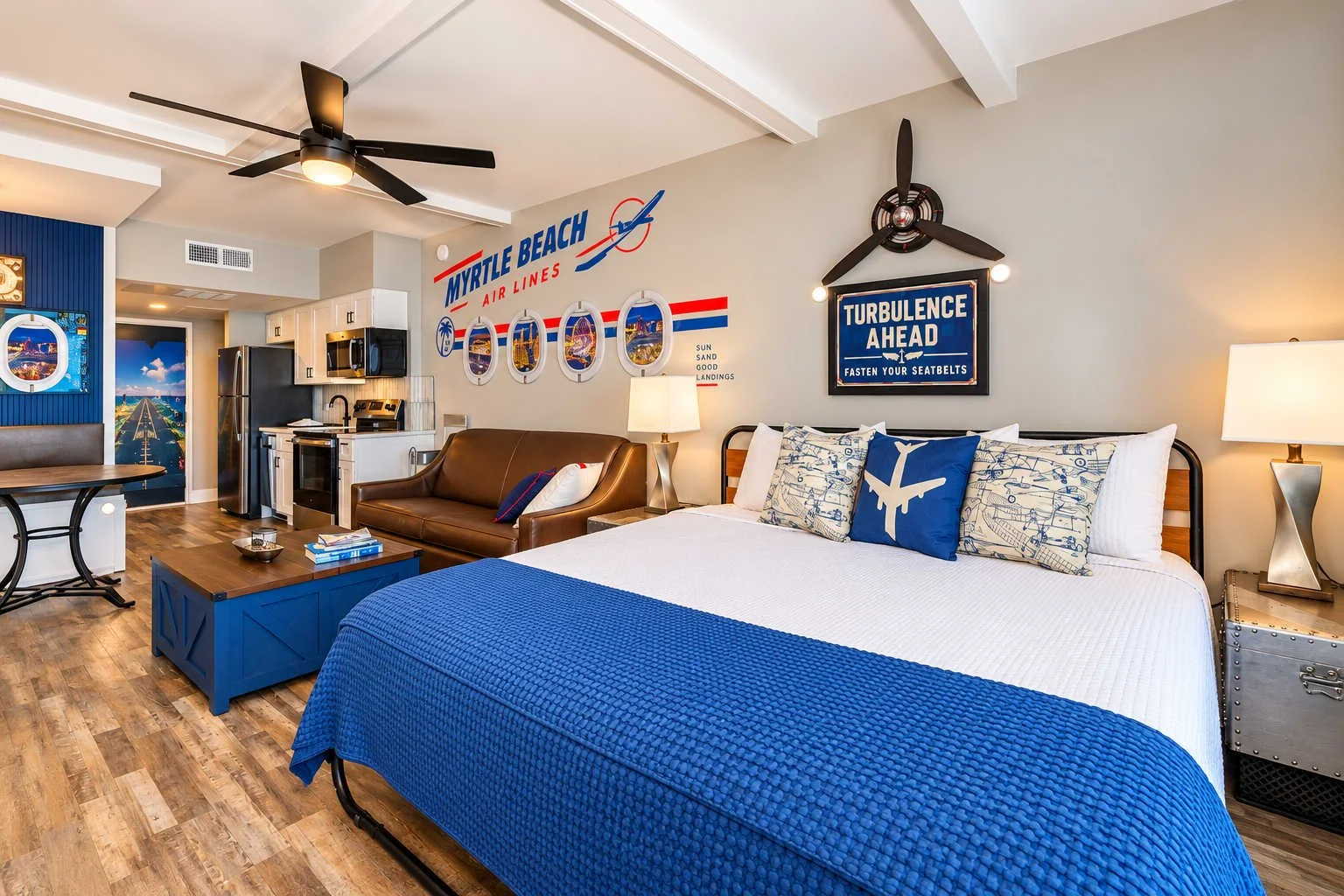 Room with a bed, pillows, and a sign on the wall that says 'Turbulence Ahead, Fasten Your Seatbelts,' decorated in a nautical and aviation theme. There is a leather sofa, a blue coffee table, a kitchenette with a refrigerator, microwave, and stove, a