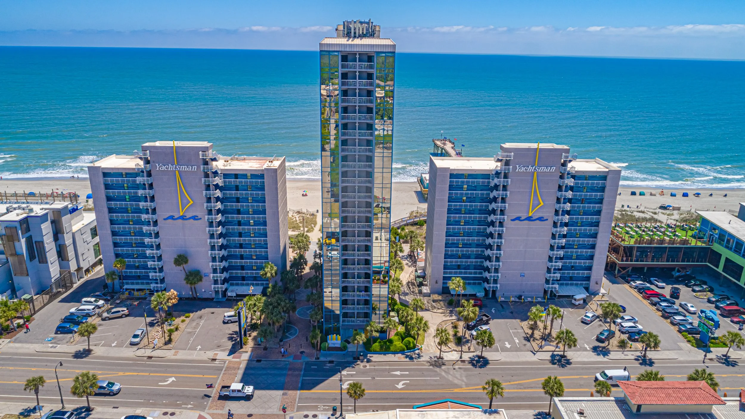 High-rise beachfront hotel named Yachtsman with a pier extending into the ocean, surrounded by a parking lot and palm trees, with the ocean and blue sky in the background.