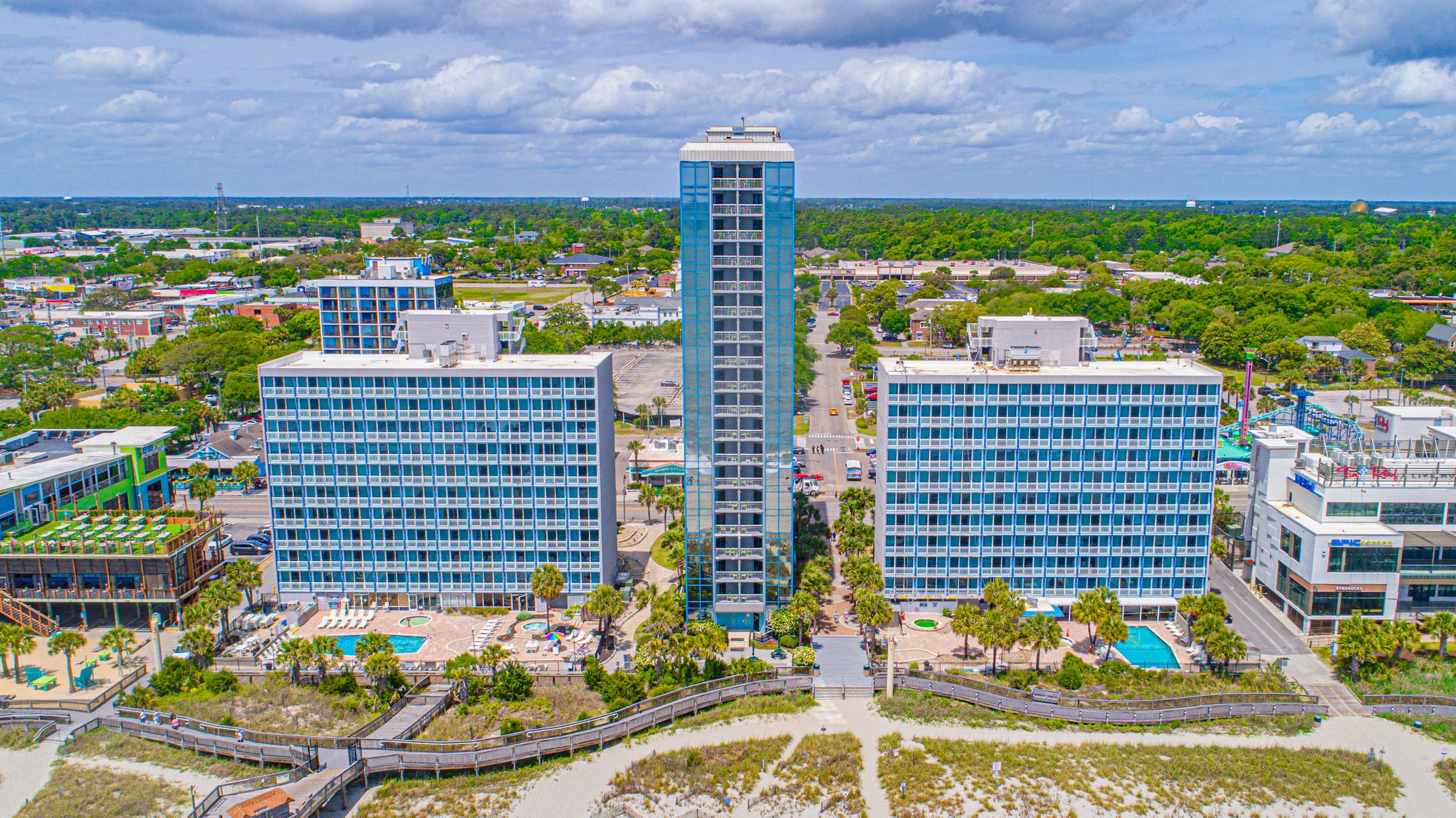 Aerial view of high-rise buildings in a city with trees, a parking lot, a swimming pool, and a beach in the foreground, under a partly cloudy sky.