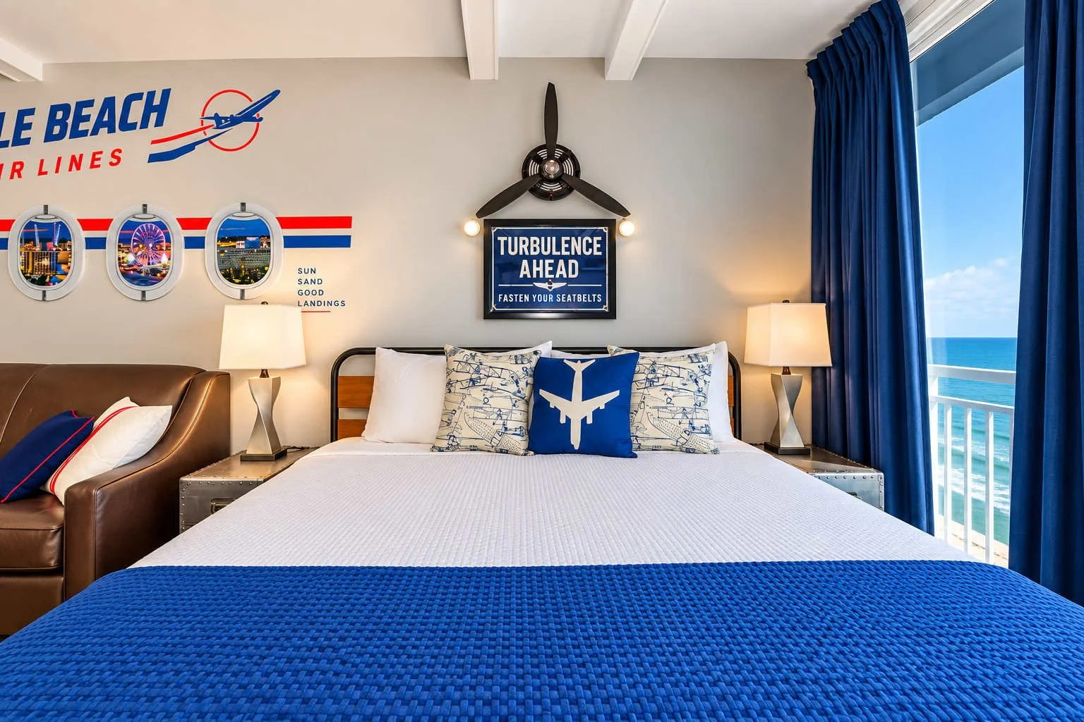 Beach-themed hotel room with airline decor, featuring a large bed with blue and white pillows, a framed sign that says 'Turbulence Ahead,' a fan with airplane blades, and ocean view through windows with blue curtains.