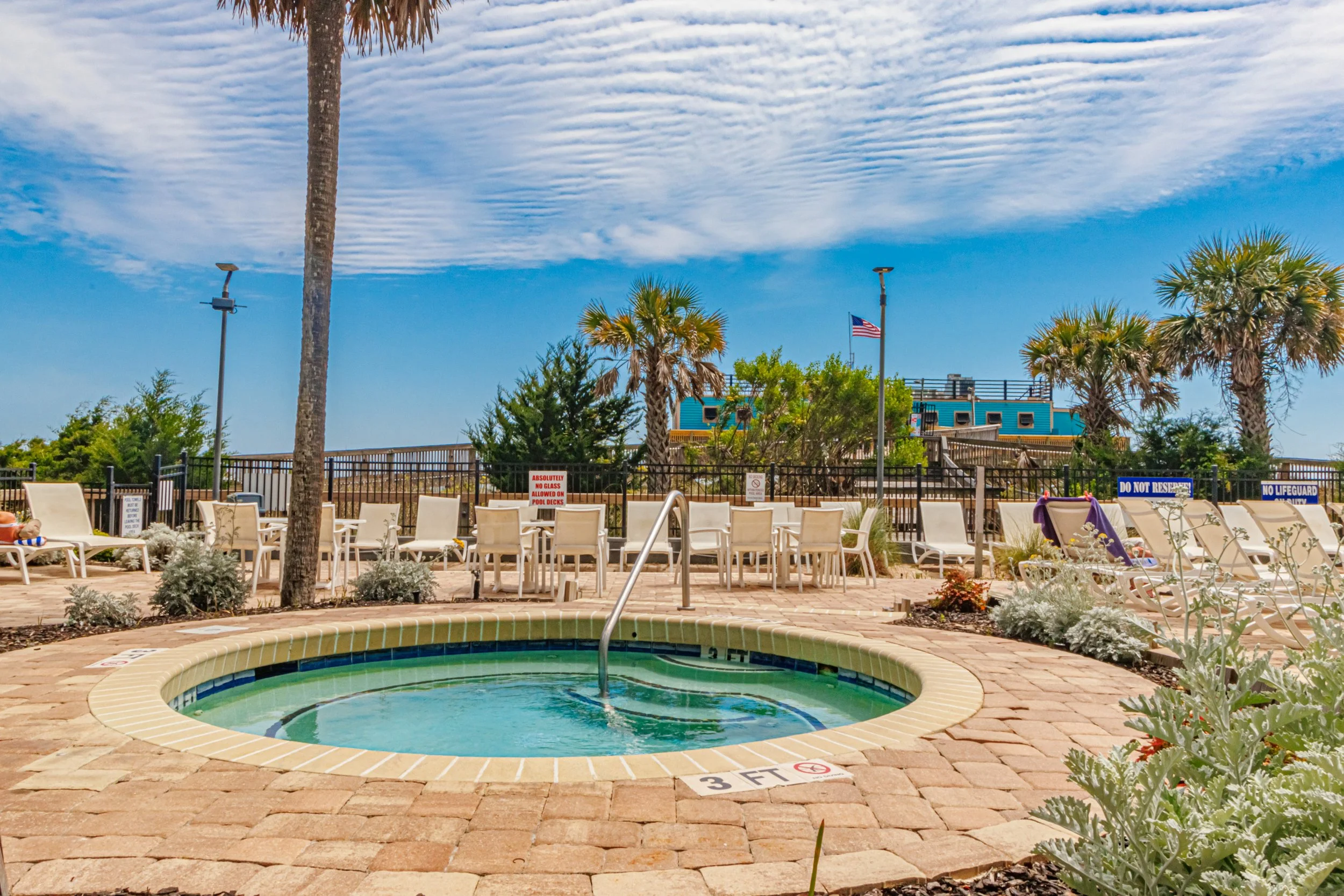 A view of a swimming pool area with lounge chairs, potted plants, and a fence with signs. Tall palm trees and a blue building with an American flag are in the background under a partly cloudy sky.