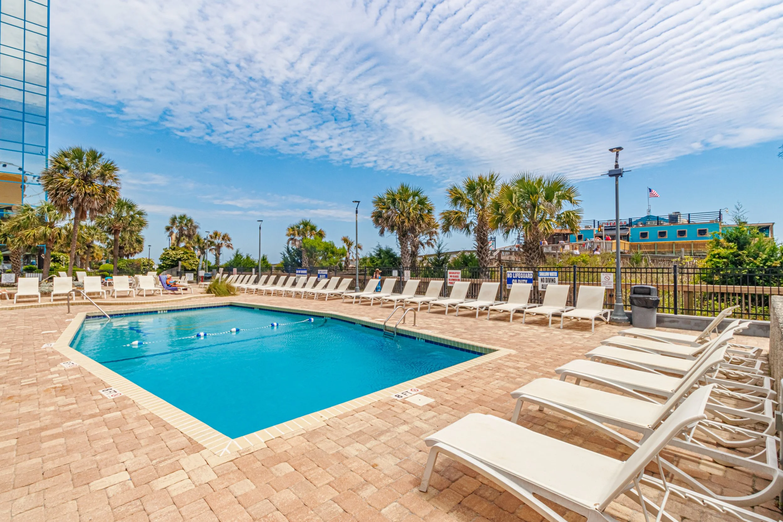 An outdoor swimming pool surrounded by white lounge chairs, with palm trees, clear sky, and a building with an American flag in the background.
