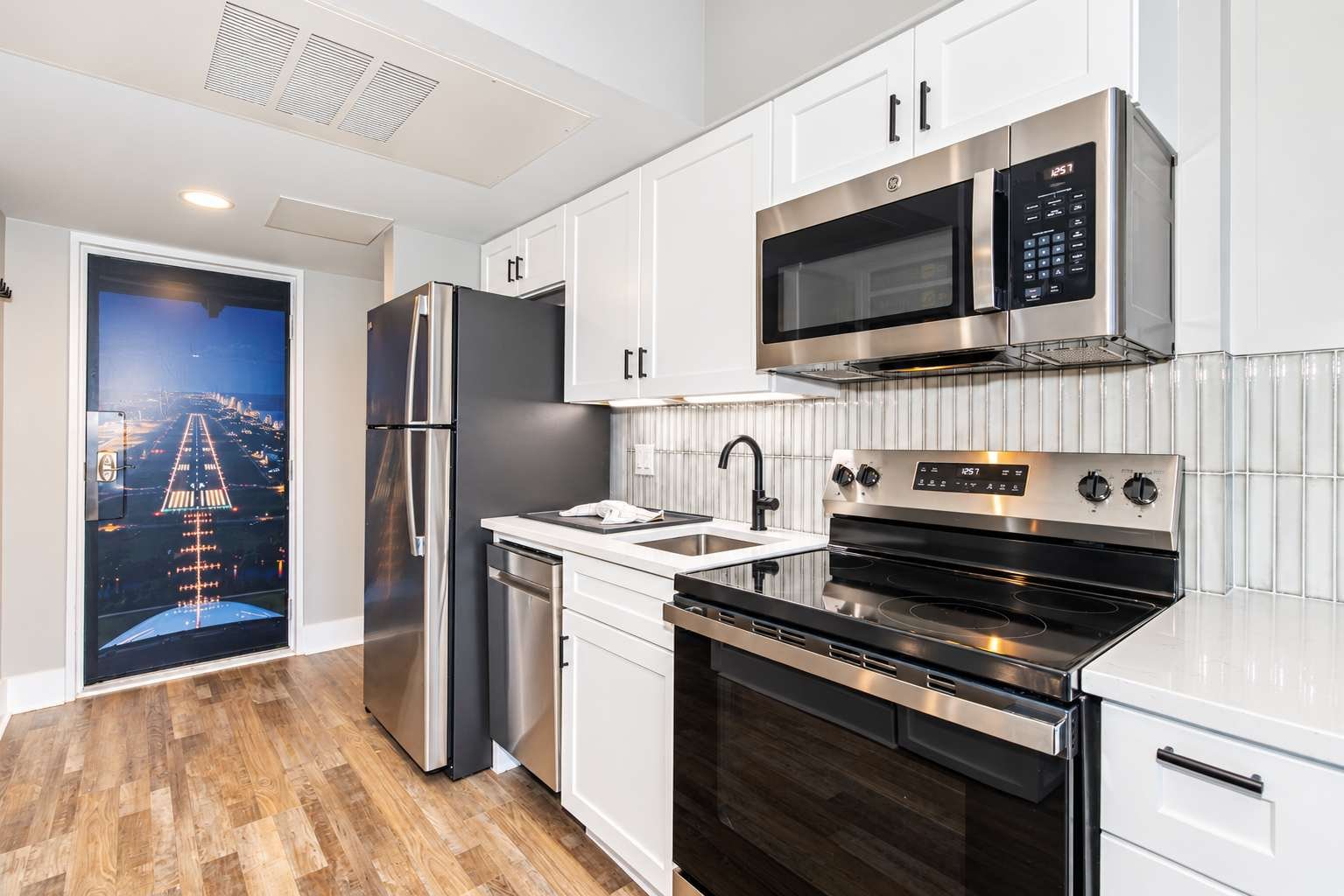 Modern kitchen with stainless steel appliances, white cabinets, a black stove, a microwave, a refrigerator, a sink with black faucet, hardwood floor, and a glass door with a view of an airport runway at night.