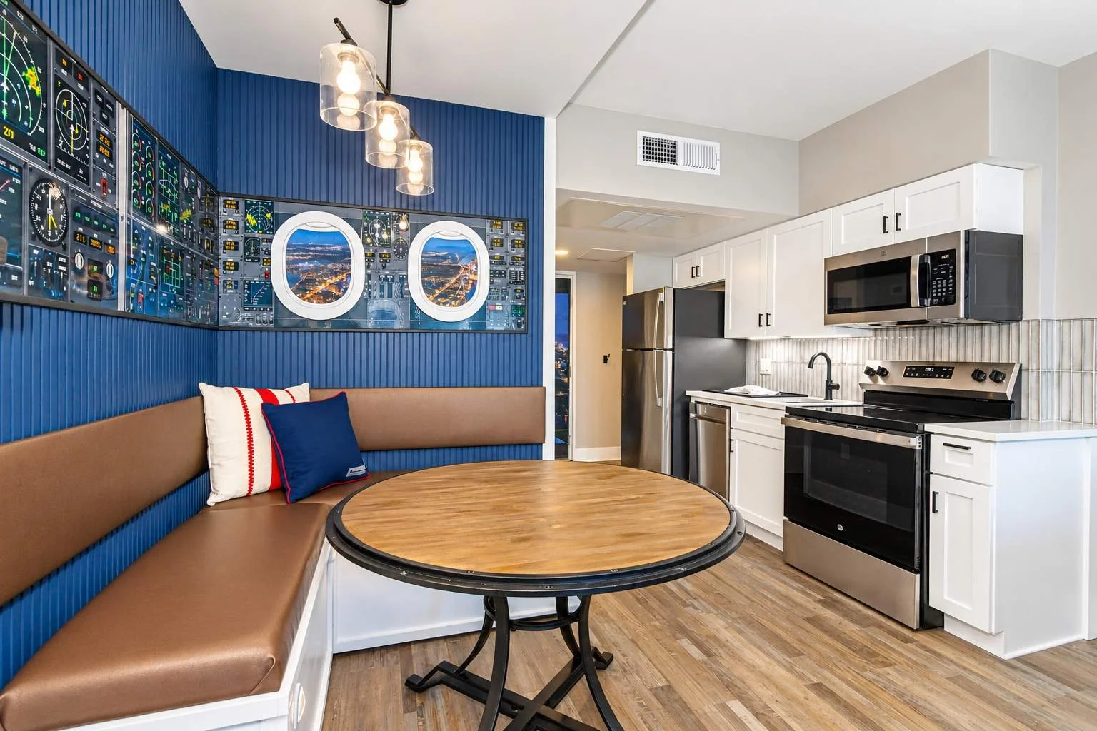 A kitchen and dining area with a blue accent wall decorated with airplane cockpit panels, a round dining table, white cabinets, stainless steel appliances, and a seating nook with pillows.
