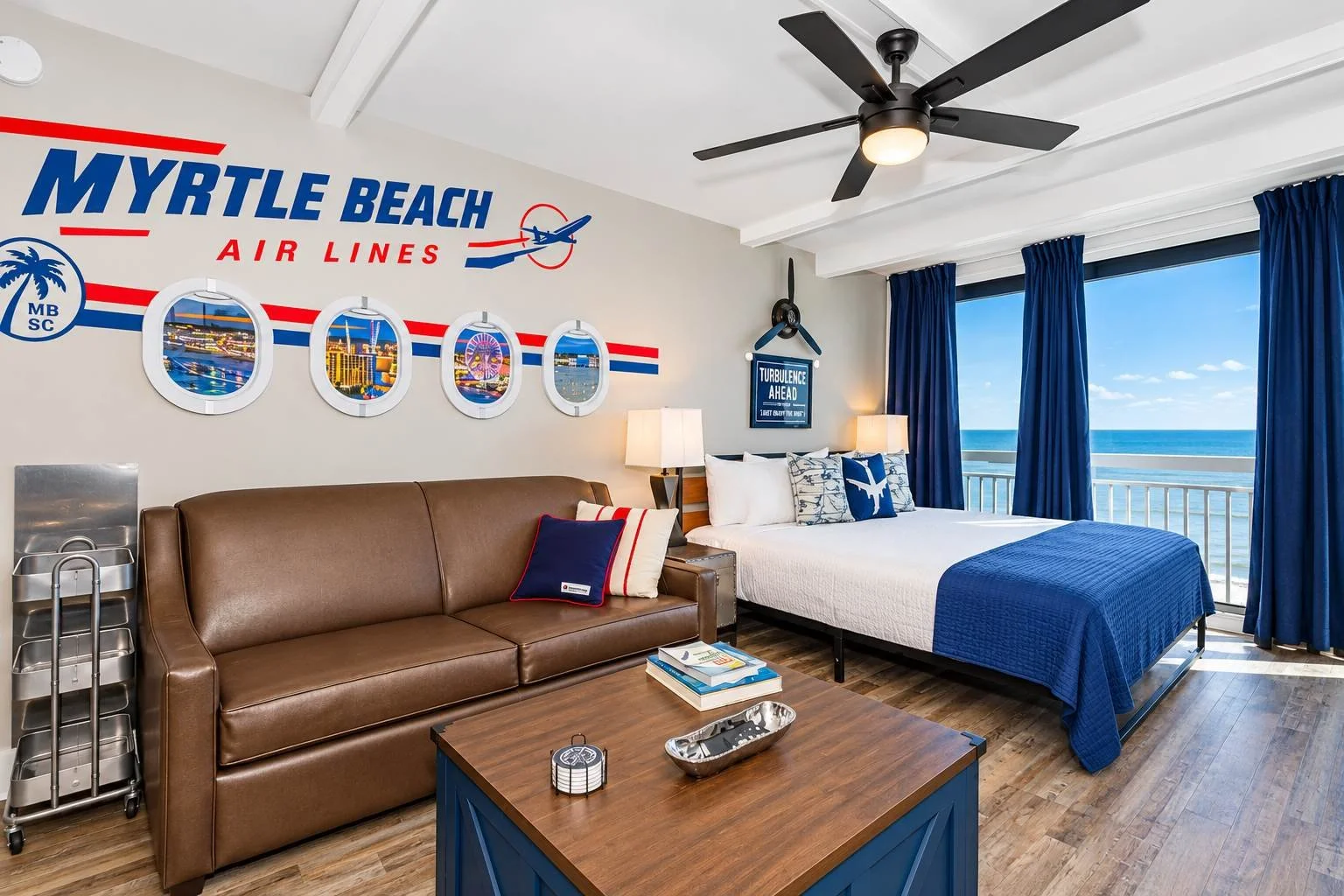 Beachside hotel room with a large window, blue curtains, and ocean view. Decor includes Myrtle Beach Air Lines wall art, a brown sofa with navy and striped pillows, a wooden coffee table with magazines and a tray, a bed with white and blue bedding, a