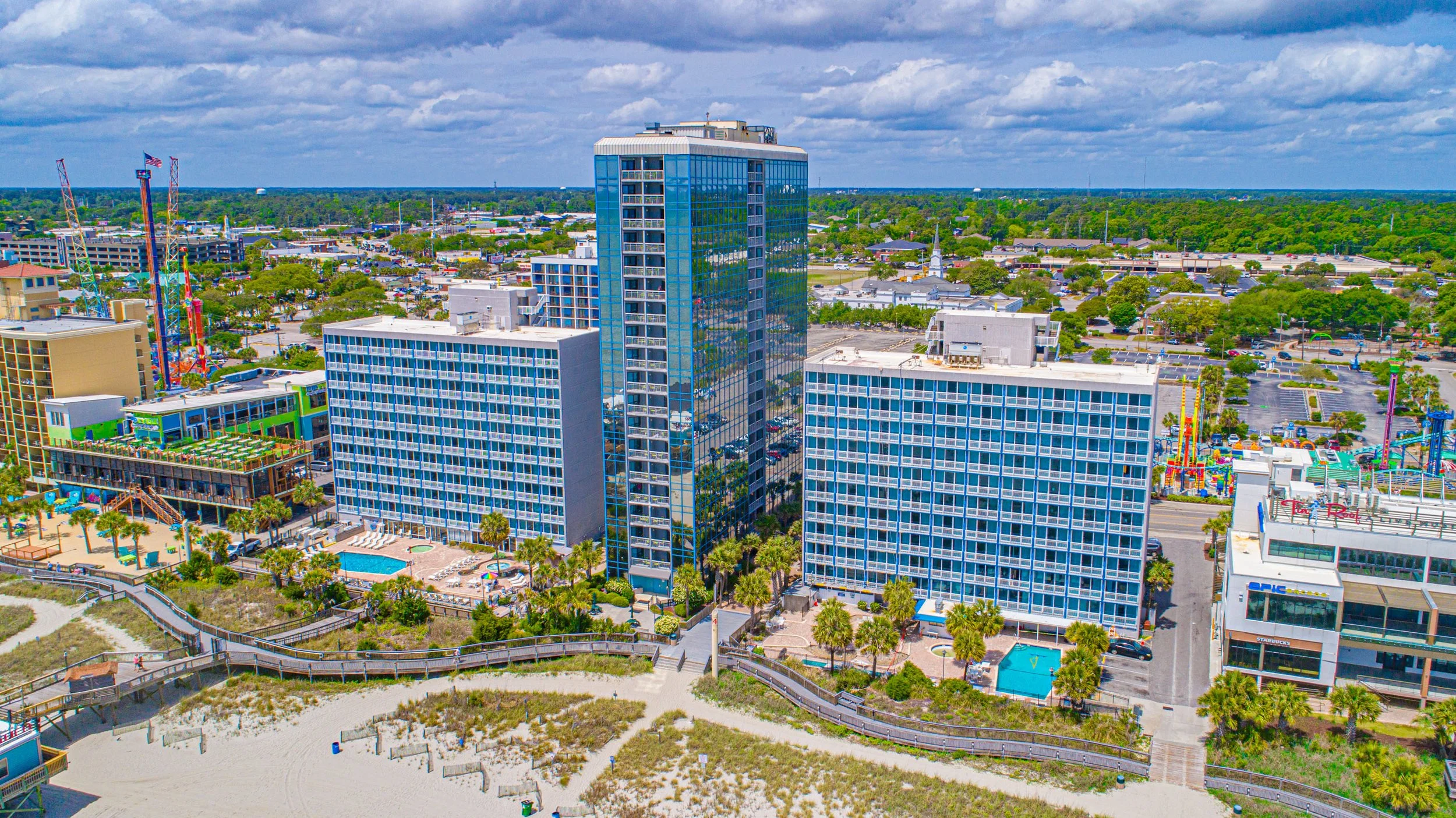 Aerial view of a cityscape with high-rise buildings, a water park, and green trees under a partly cloudy sky.
