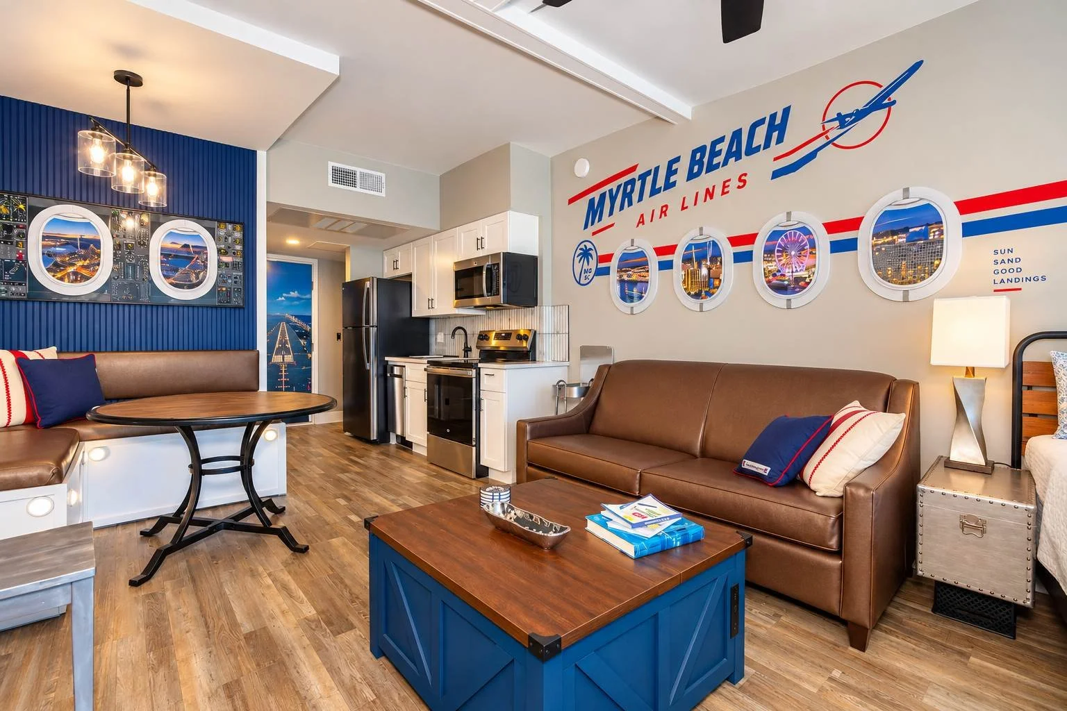 Living room with nautical beach theme decor, featuring a brown leather sofa, a blue coffee table, a small dining area, a kitchenette, and wall art themed around Myrtle Beach air lines and aviation, with a color scheme of blue, red, and white.