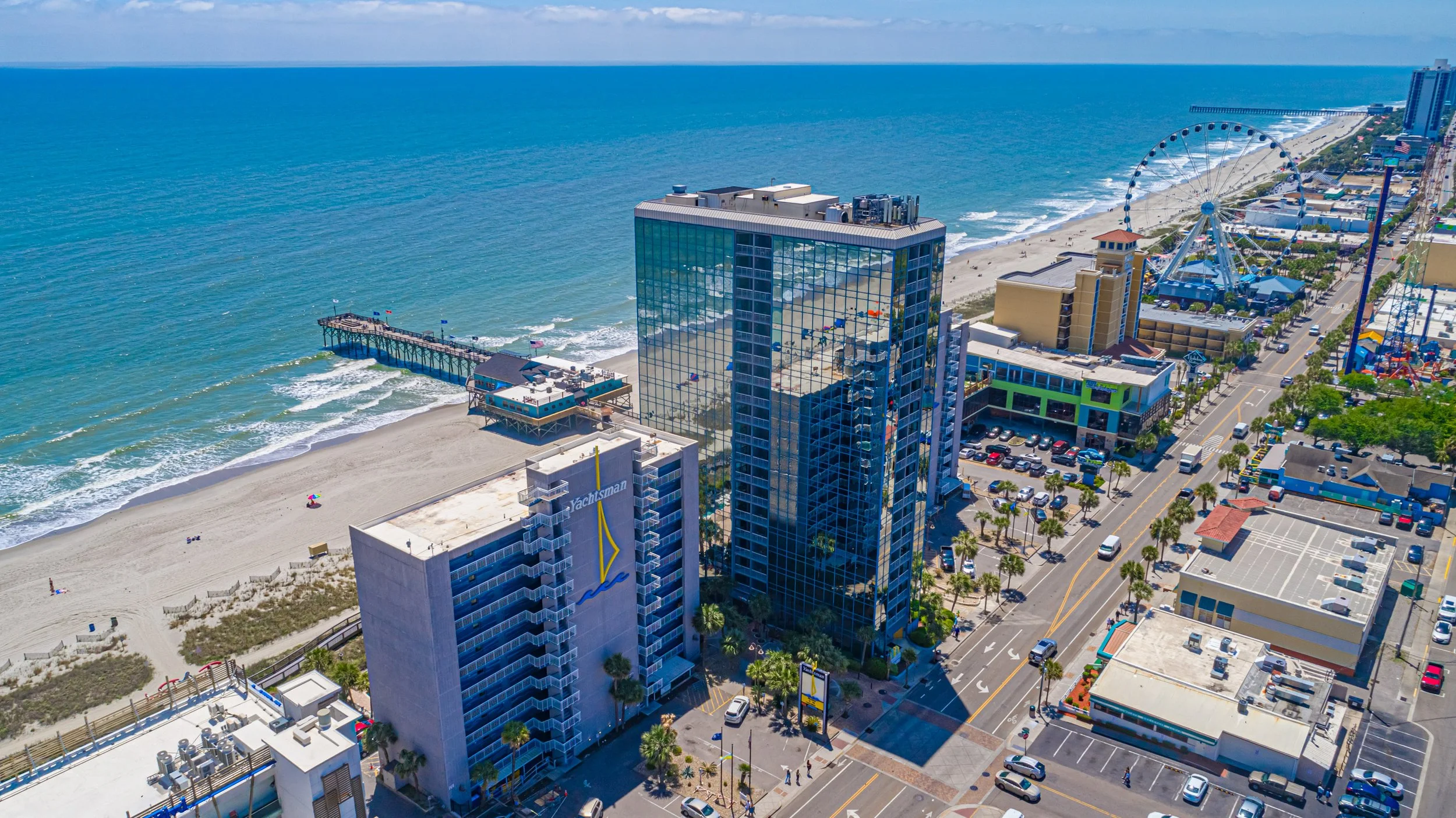 Aerial view of a beachside city with high-rise buildings, a pier extending into the ocean, a Ferris wheel, and a busy street lined with palm trees.