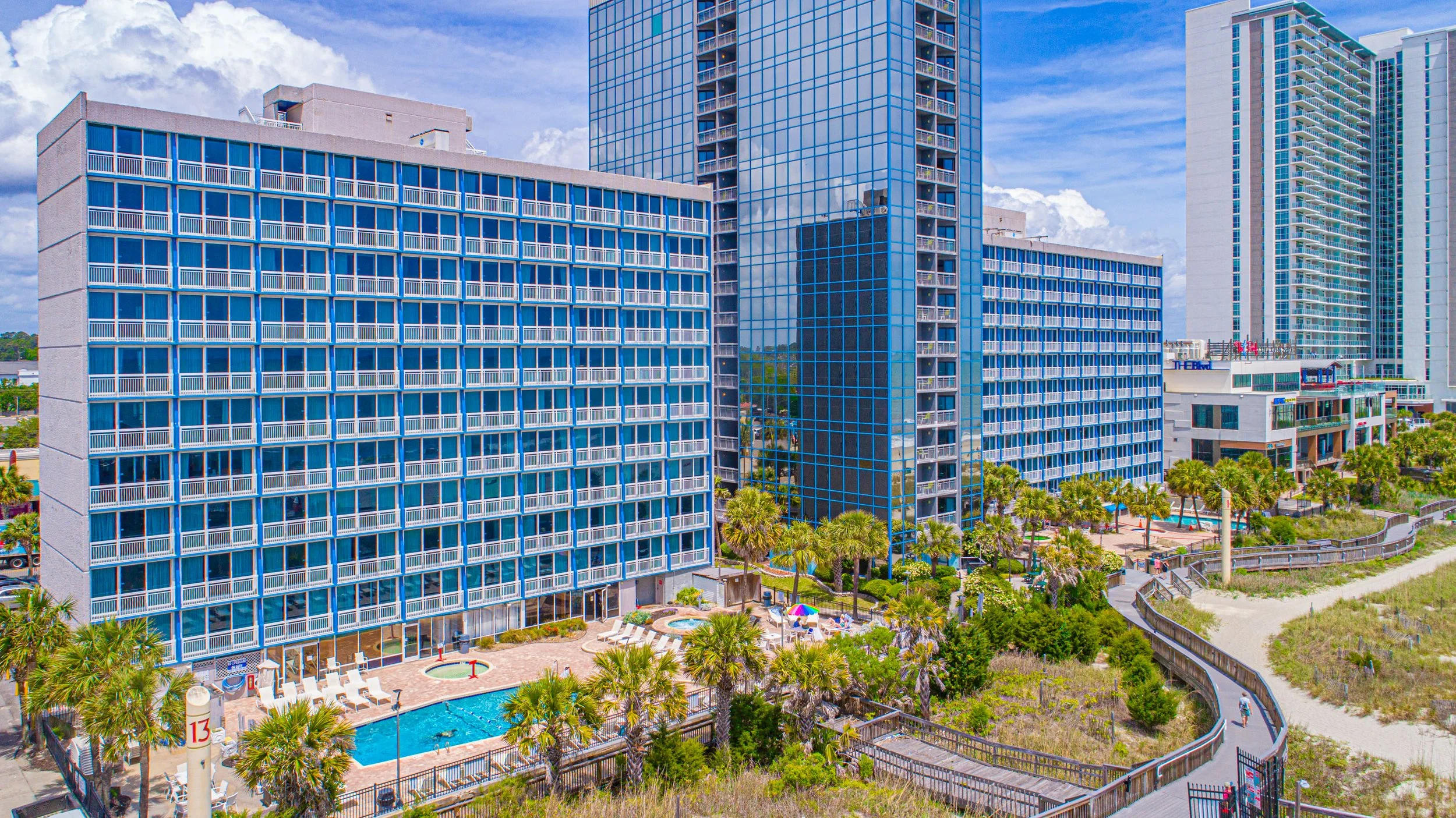 High-rise hotel buildings near a swimming pool with lounge chairs, surrounded by palm trees and greenery, under a partly cloudy sky.