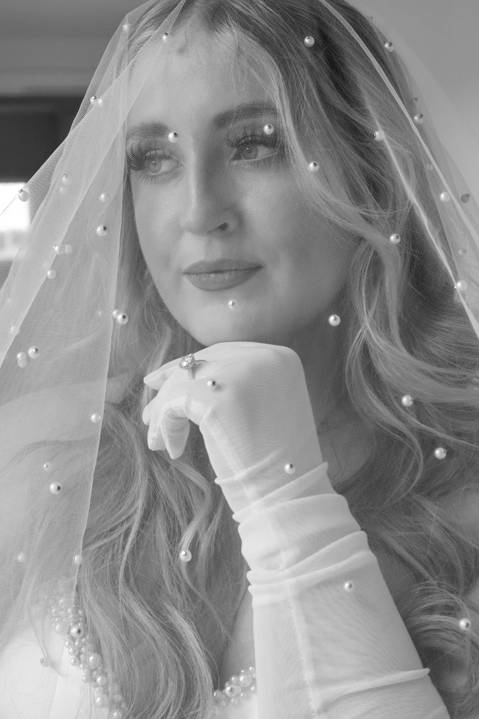 A woman with long curled hair, wearing a wedding veil with pearls, a ring, and white gloves, looking thoughtfully with her chin resting on her hand.