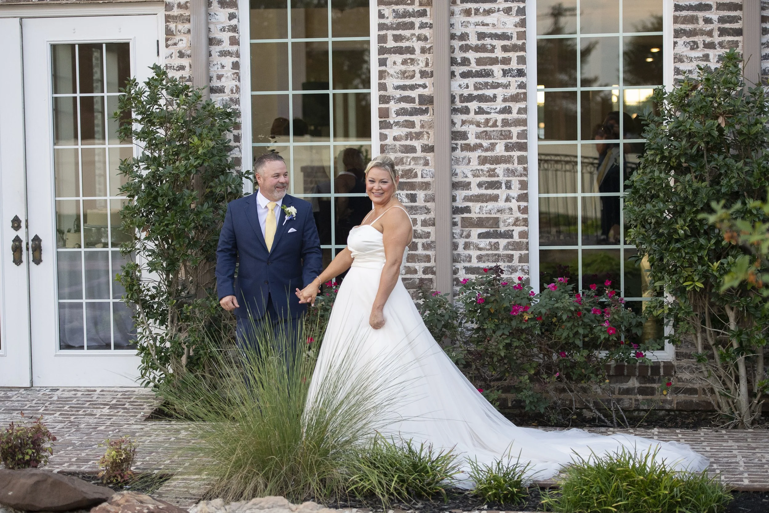 A bride and groom holding hands outside a brick house with large windows. The bride is wearing a white wedding dress, and the groom is dressed in a navy blue suit with a yellow tie. They are smiling and looking at each other, surrounded by greenery a