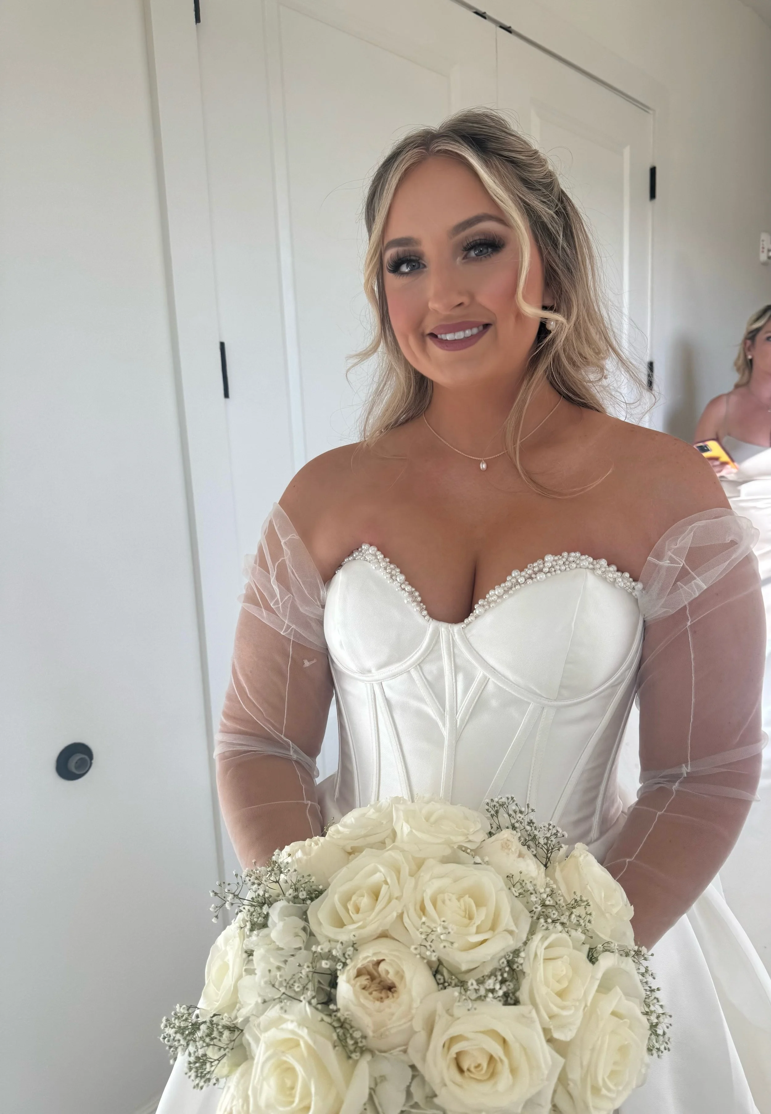 A smiling bride in a white wedding dress holding a bouquet of white roses, standing indoors.