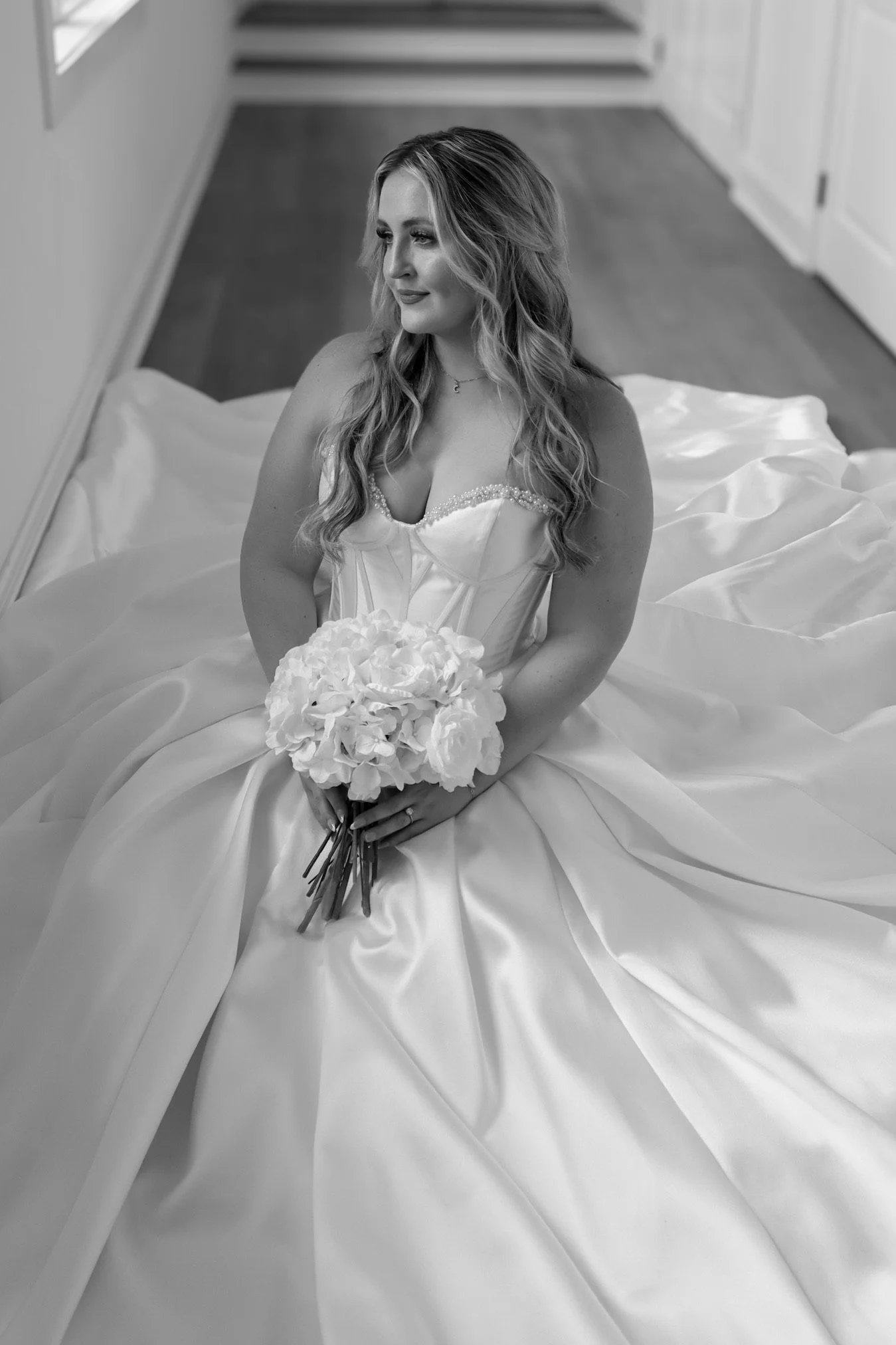 Black and white photo of a bride in a wedding dress, sitting on a bed, holding a bouquet of flowers, and looking to the side.