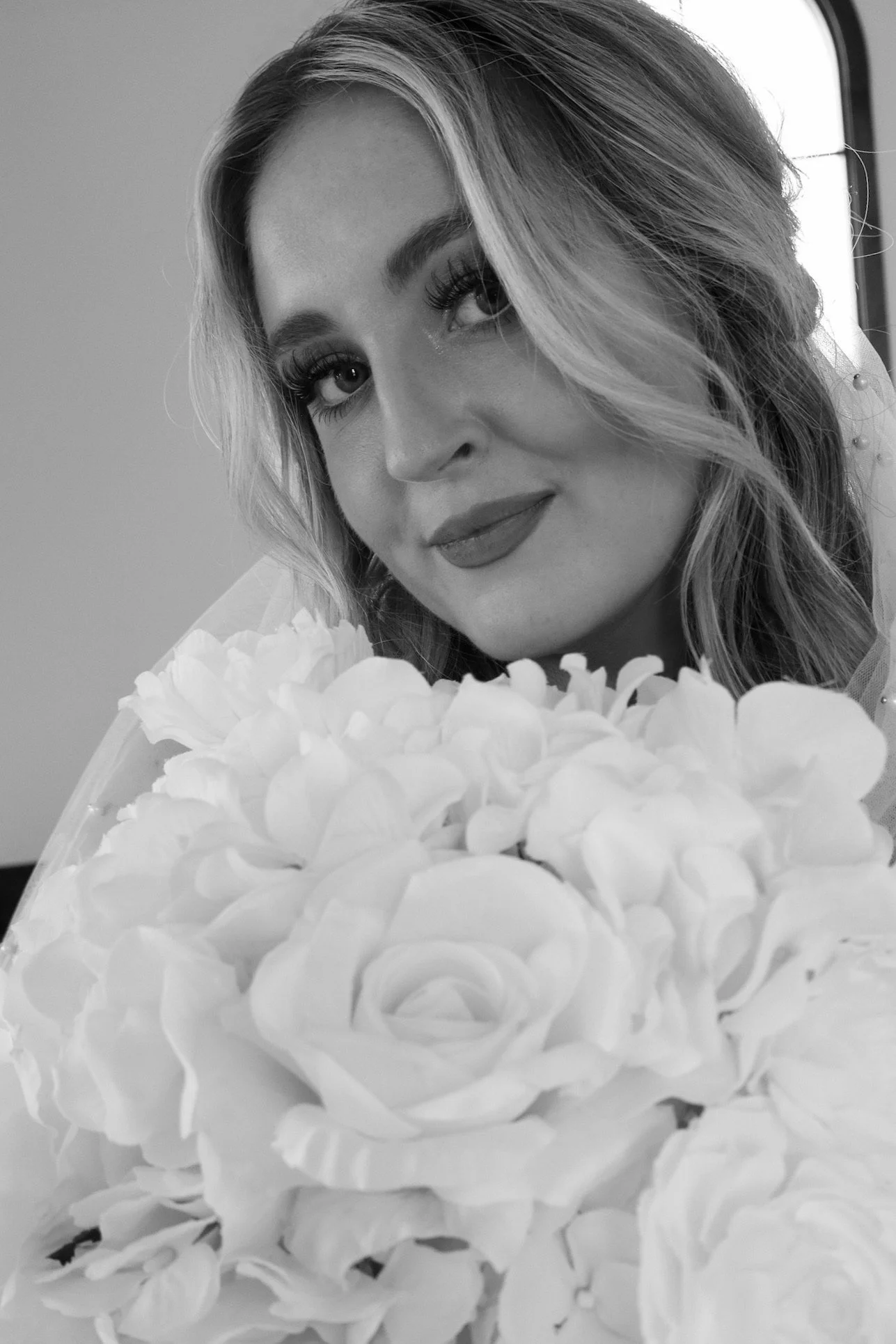 A woman with wavy blonde hair and makeup holding a large bouquet of flowers, smiling softly.