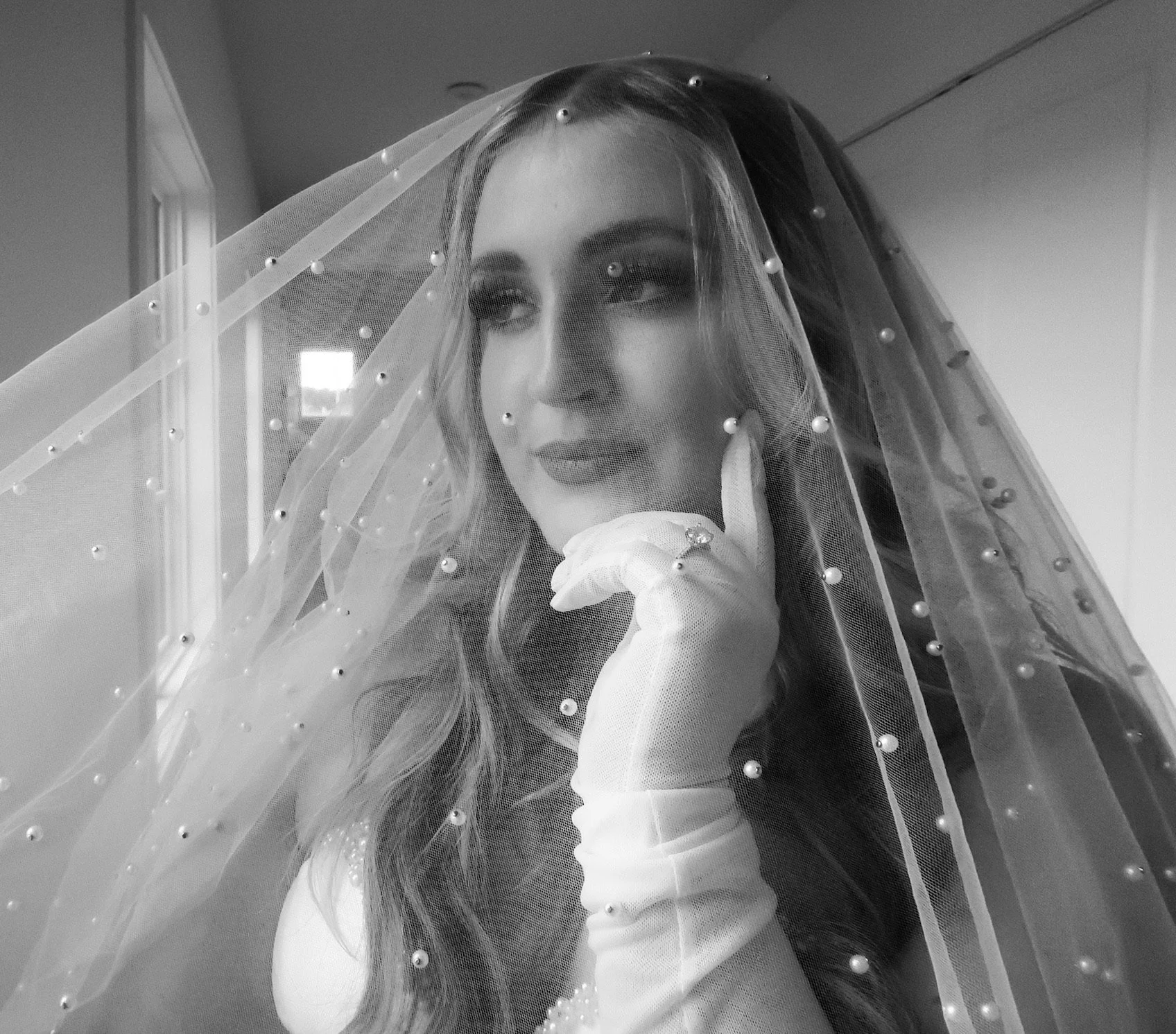 A woman wearing a wedding veil and an elegant dress, with a thoughtful expression, under natural light coming from a window.