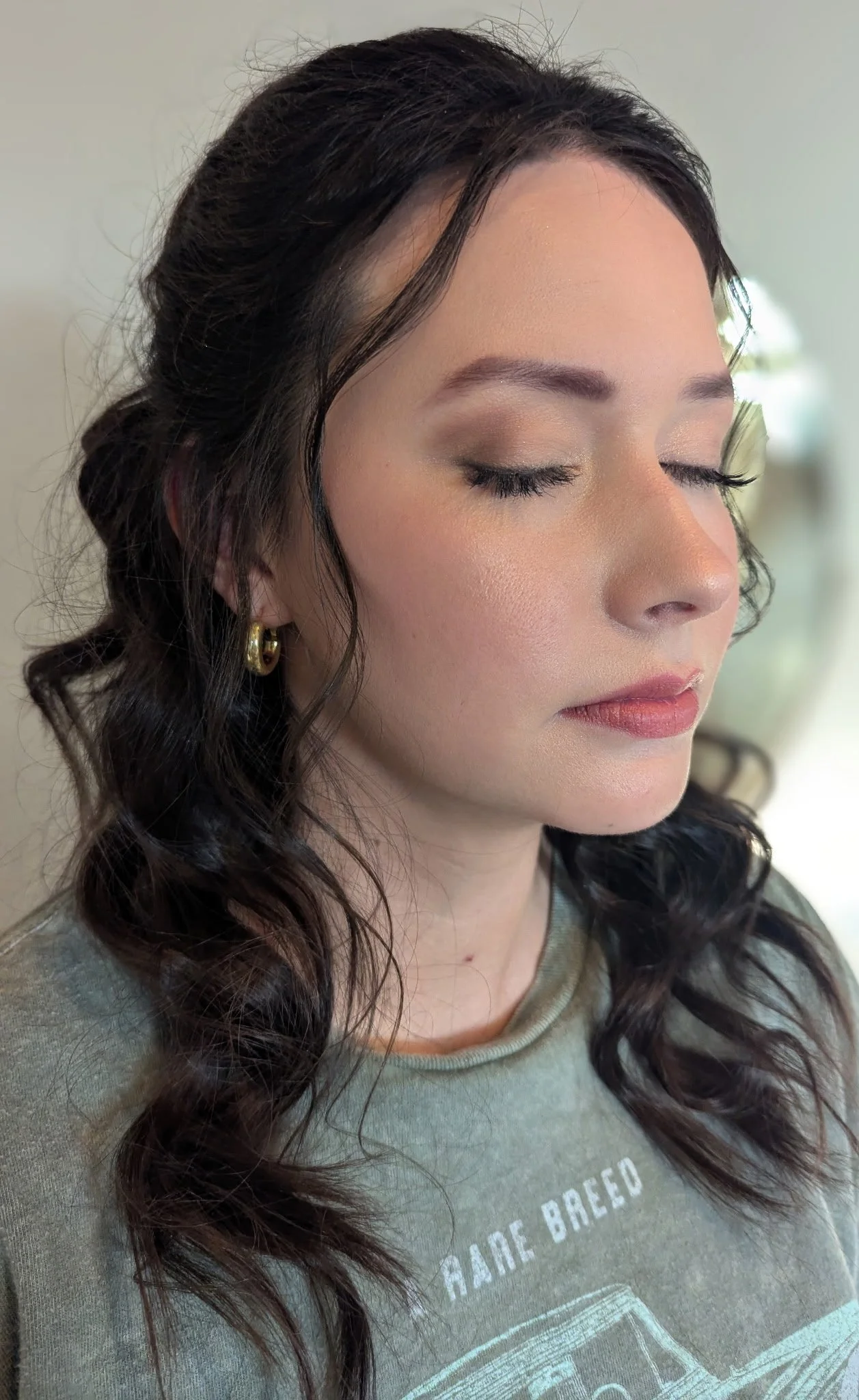 Close-up of a young woman with dark, wavy hair and light makeup with eyes closed, wearing gold hoop earrings and a green shirt.