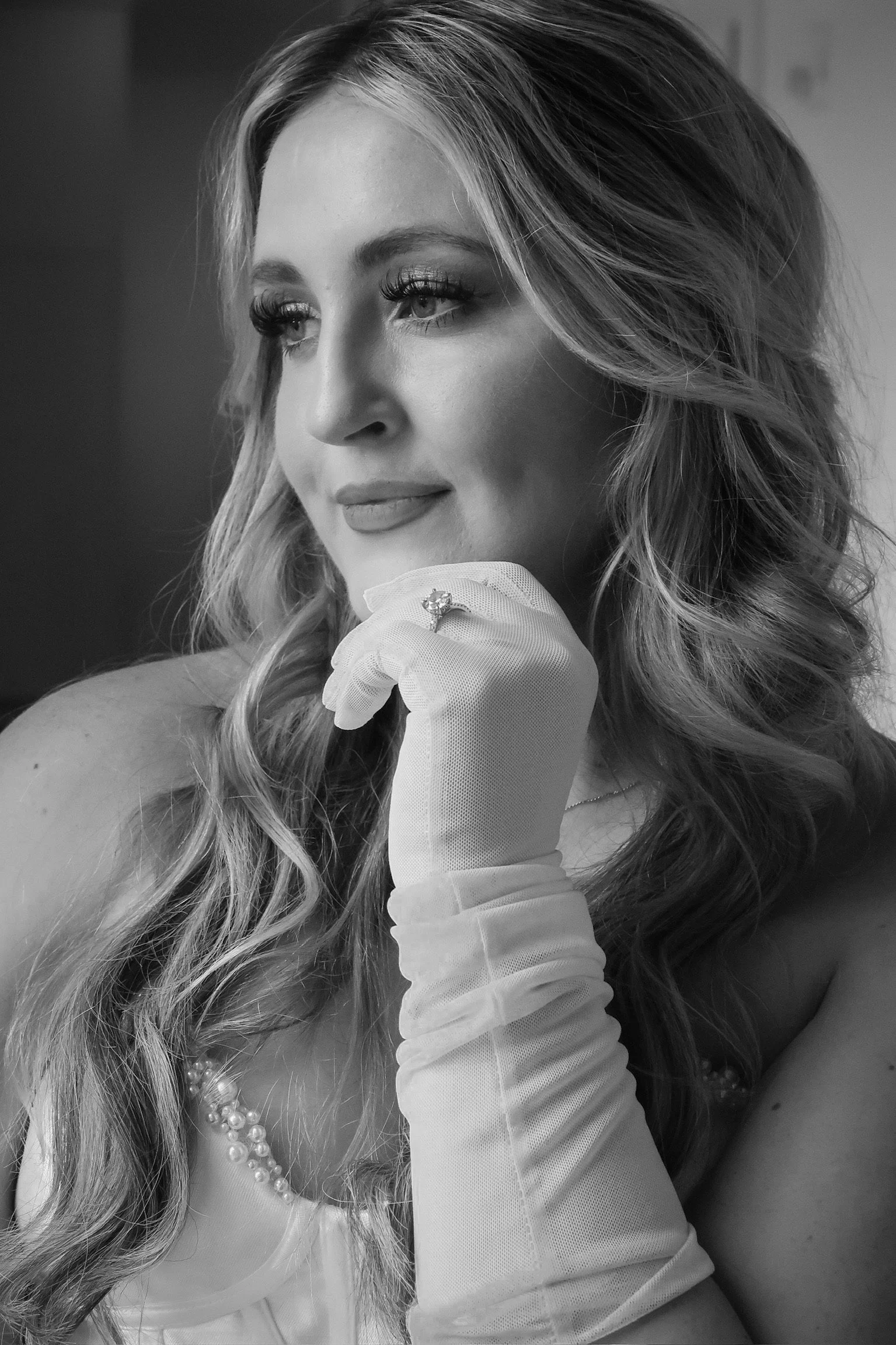 A woman with long, curly hair wearing a pearl necklace and an elegant dress, resting her chin on her hand with a ring on her finger, looking thoughtfully to the side.