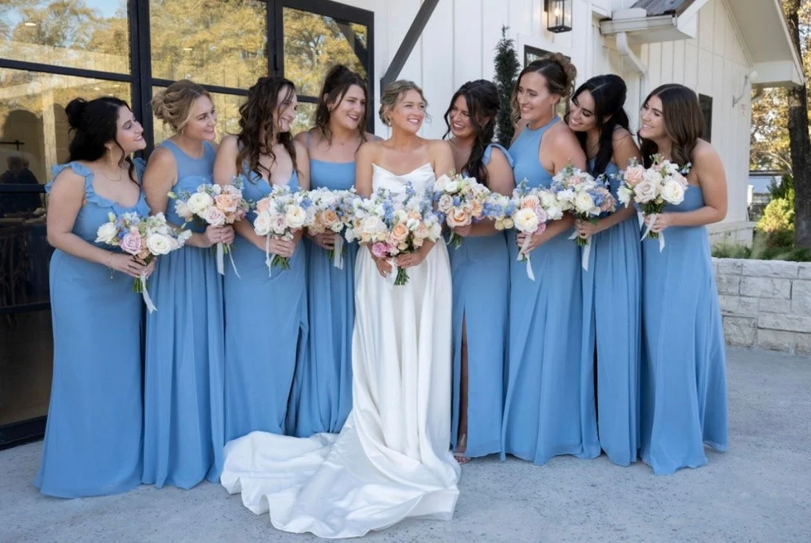 Bride and eight bridesmaids standing outside, all holding bouquets. The bride is in a white gown, while the bridesmaids are in matching blue dresses. They are smiling and looking at each other.