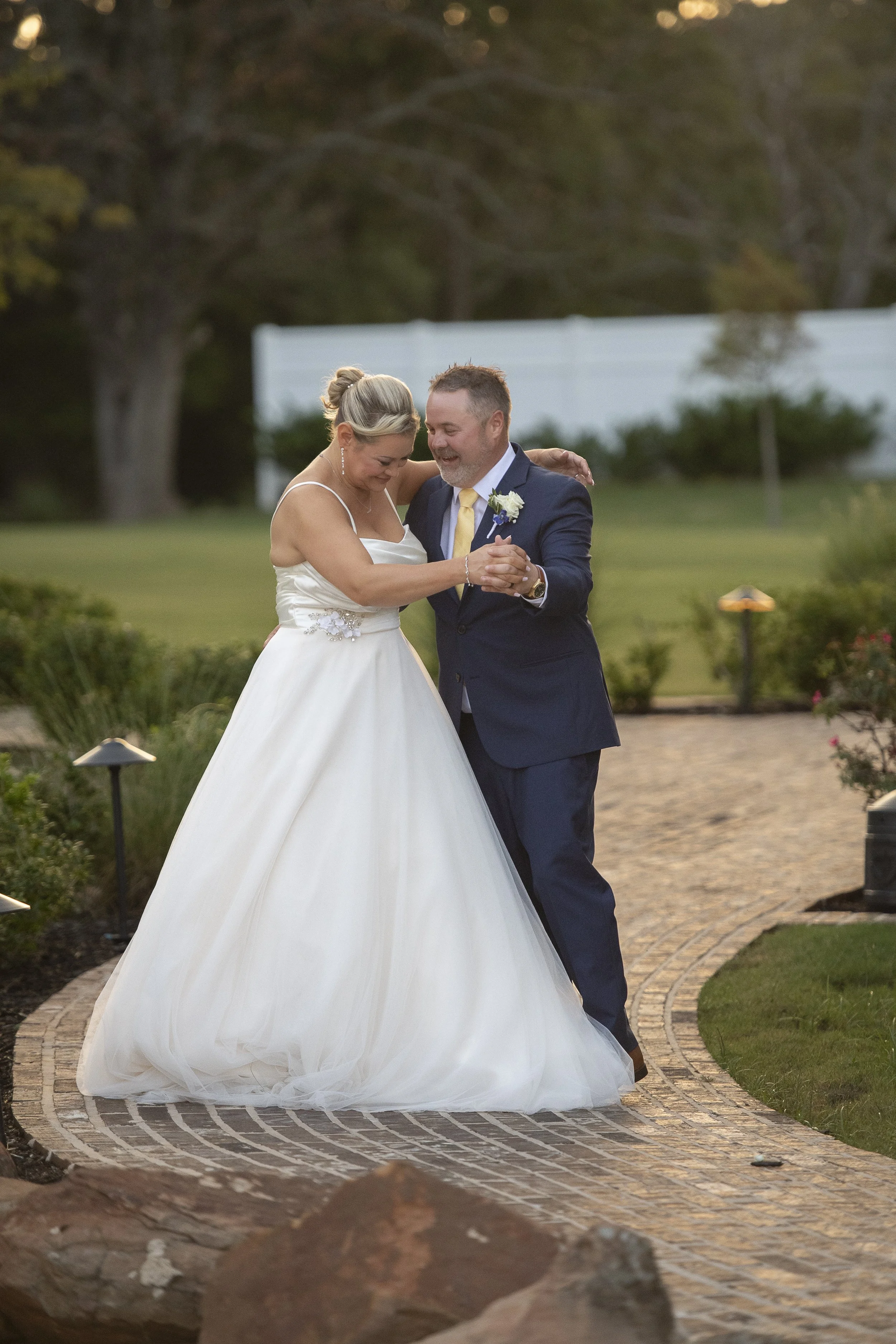 A bride and groom dancing outdoors on a brick pathway, surrounded by greenery and garden lights during sunset.