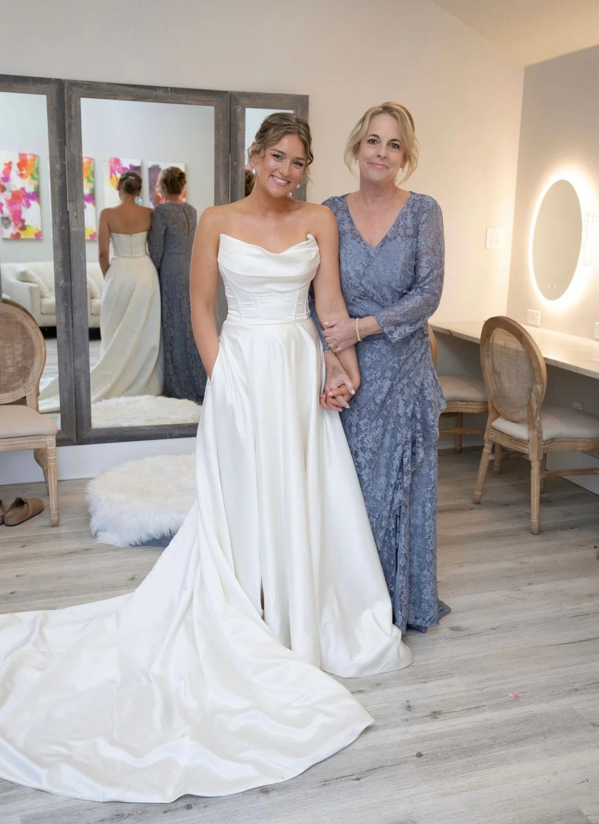 A young woman in a white wedding dress and an older woman in a blue lace dress standing together in a dressing room, holding hands and smiling.