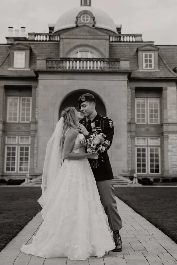 A bride and a groom in a military uniform sharing a kiss outside a grand building, with the bride holding a bouquet, in black and white.