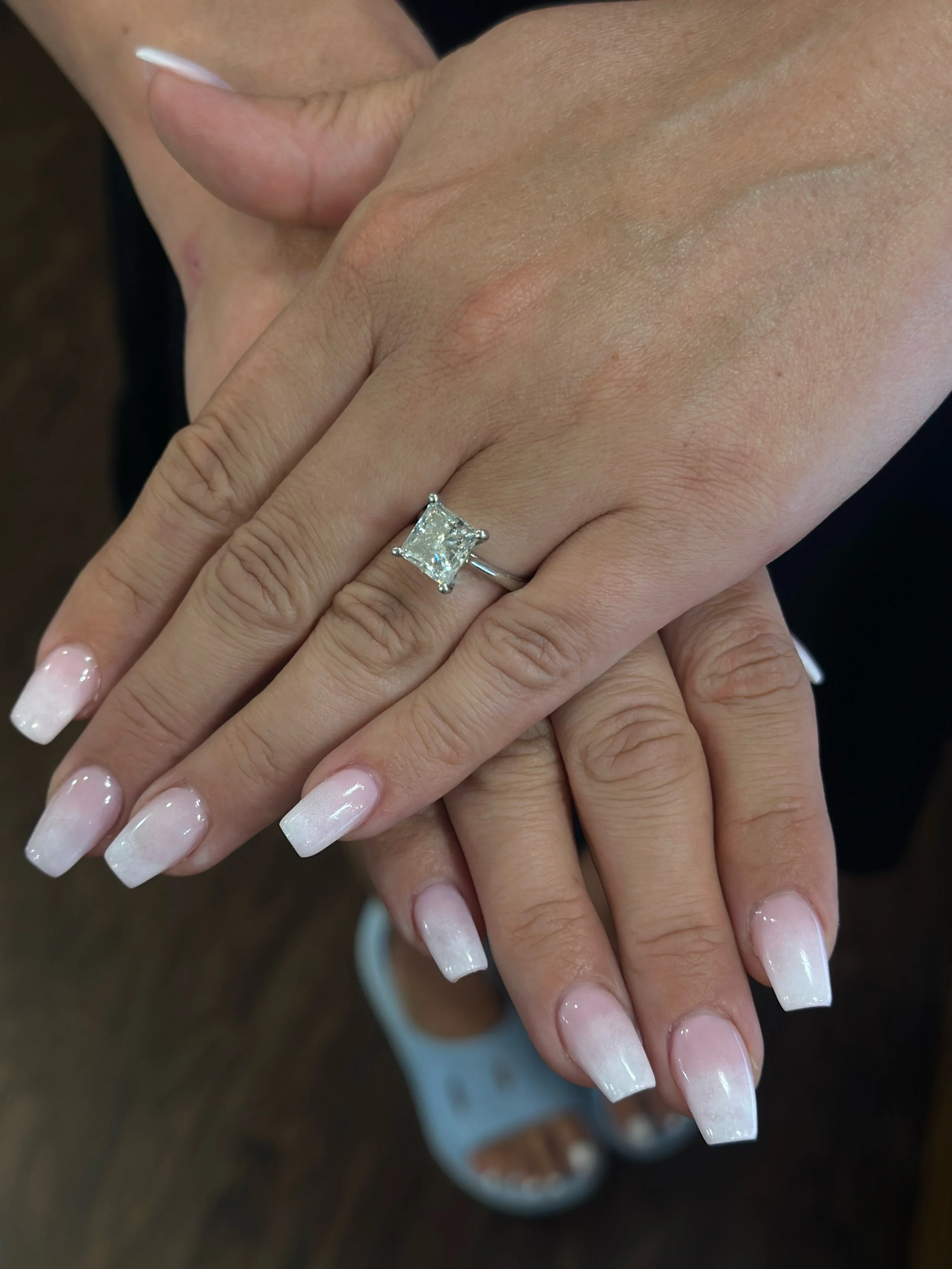 Close-up of a woman’s hands displaying a large engagement ring with a square-cut diamond on her left ring finger. She has French manicure nails with white tips and a clear shine. The background is blurred, showing part of her outfit and a wooden surf