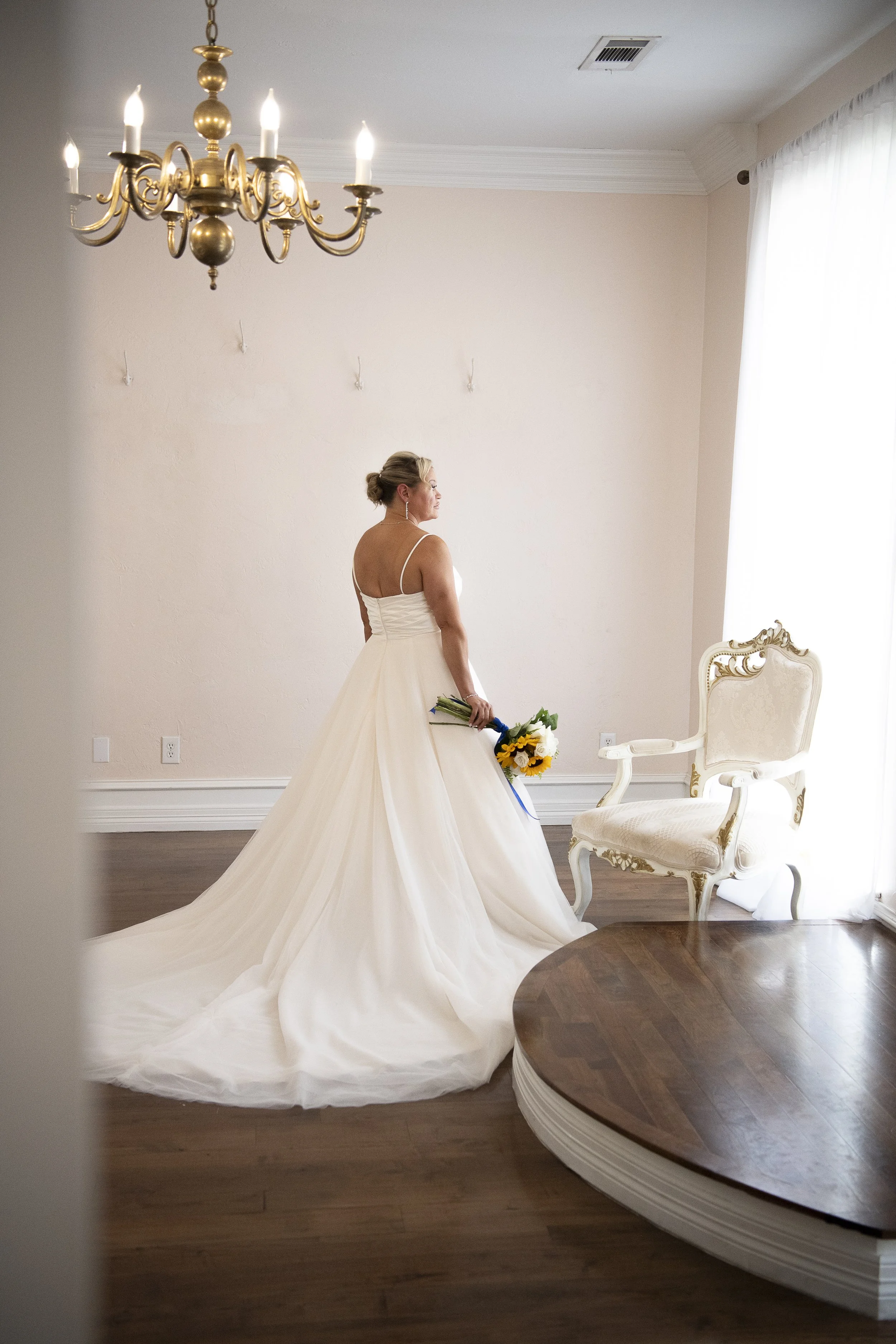A bride in a white wedding gown holding a colorful bouquet, standing in a room with hardwood floors, a vintage armchair, and a chandelier, gazing out of a window.