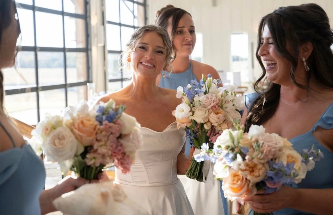 A bride holding a bouquet of flowers, smiling at bridesmaids who are also holding bouquets, inside a bright room with large windows.