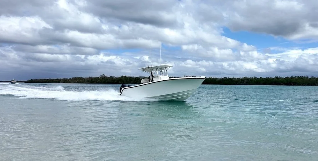 A white motorboat speeding across turquoise water with a trail of white foam, under cloudy skies with some blue visible, and a distant land covered with trees in the background.