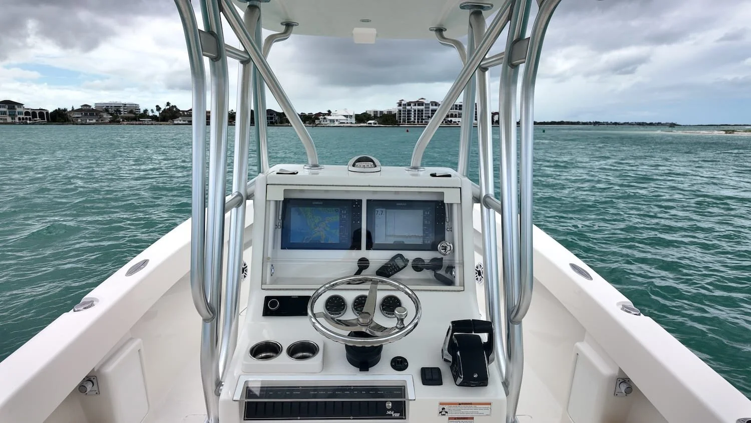 View from the helm of a white motorboat with navigation screens, steering wheel, and controls, on calm water with a cloudy sky and distant buildings.