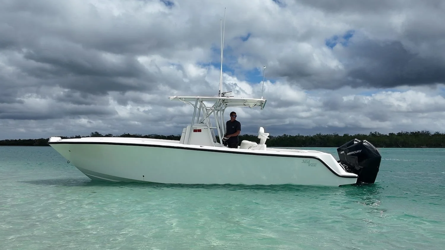 A white boat with a black stripe floats on calm, turquoise water under a cloudy sky. A man stands in the center, near the console, with a black shirt and sunglasses.