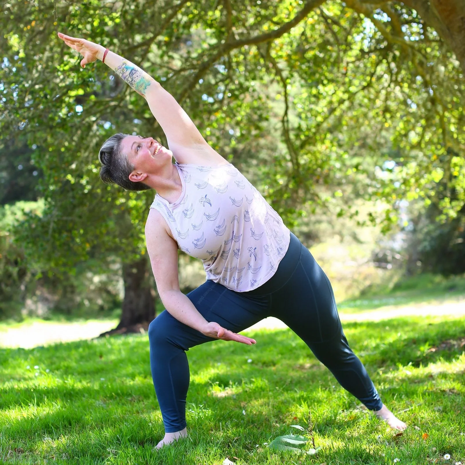Liz is in a deep lunge, her feet in green grass, summer time trees behind her. She is wearing dark blue leggings and a light purple tank top with a print of bananas in blue wearing glasses. One of her arms is bent on her leg, the other arches over.